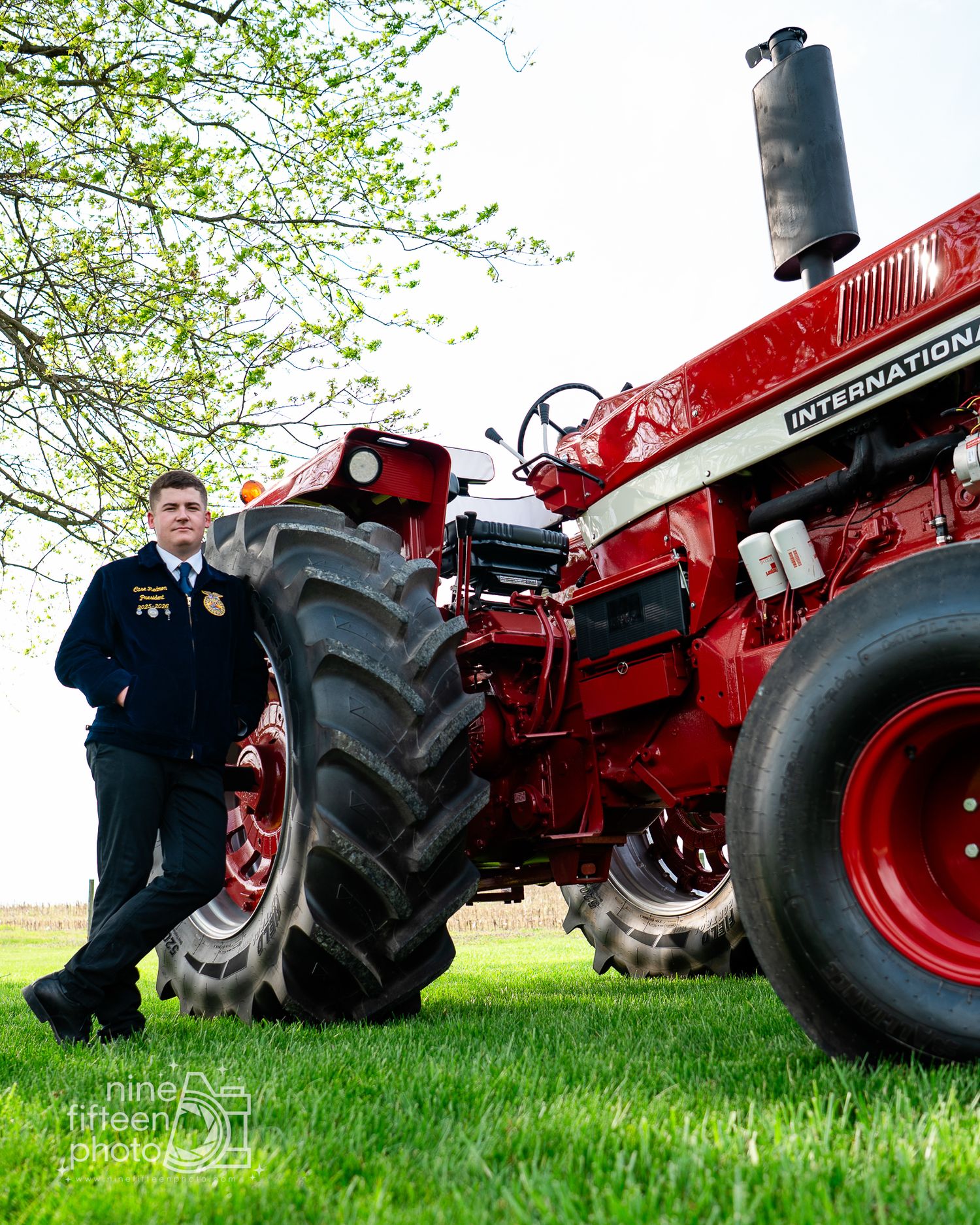 FFA member in blue jacket poses beside a classic red International tractor on green grass under a tree.