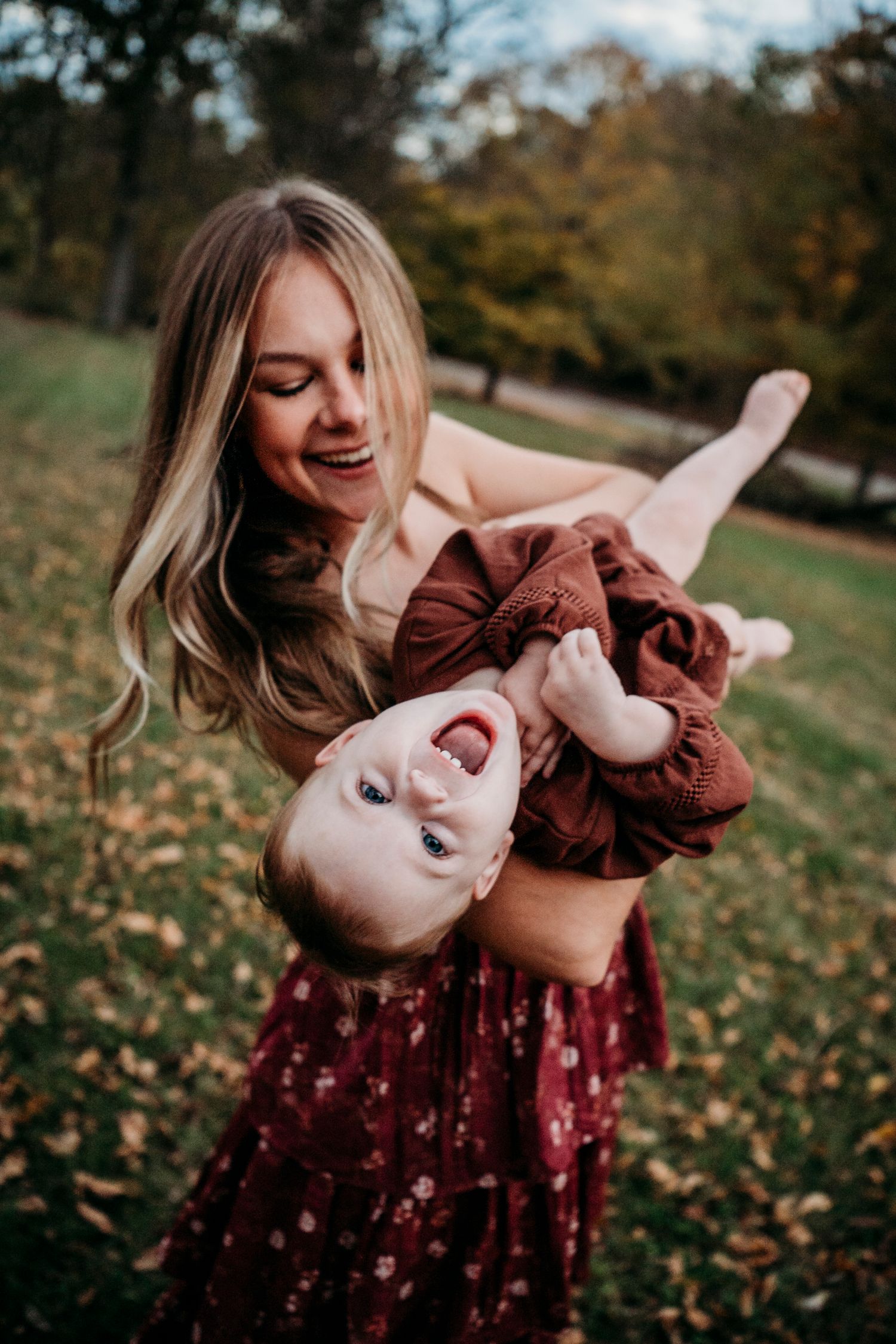 A joyful mother holds her laughing baby upside down outdoors in an autumn park, both wearing burgundy outfits.