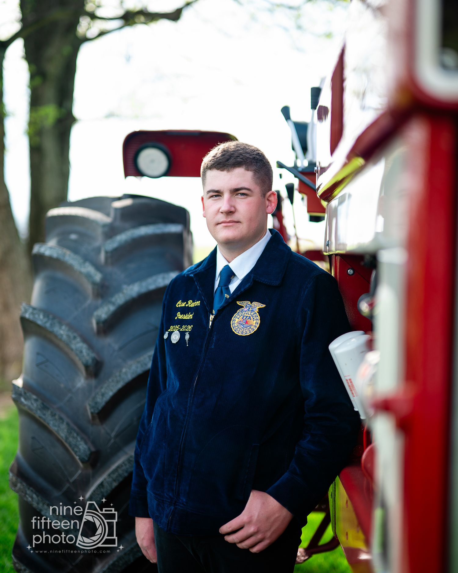Young man in FFA jacket stands confidently beside a red tractor outdoors in a professional senior portrait photo.