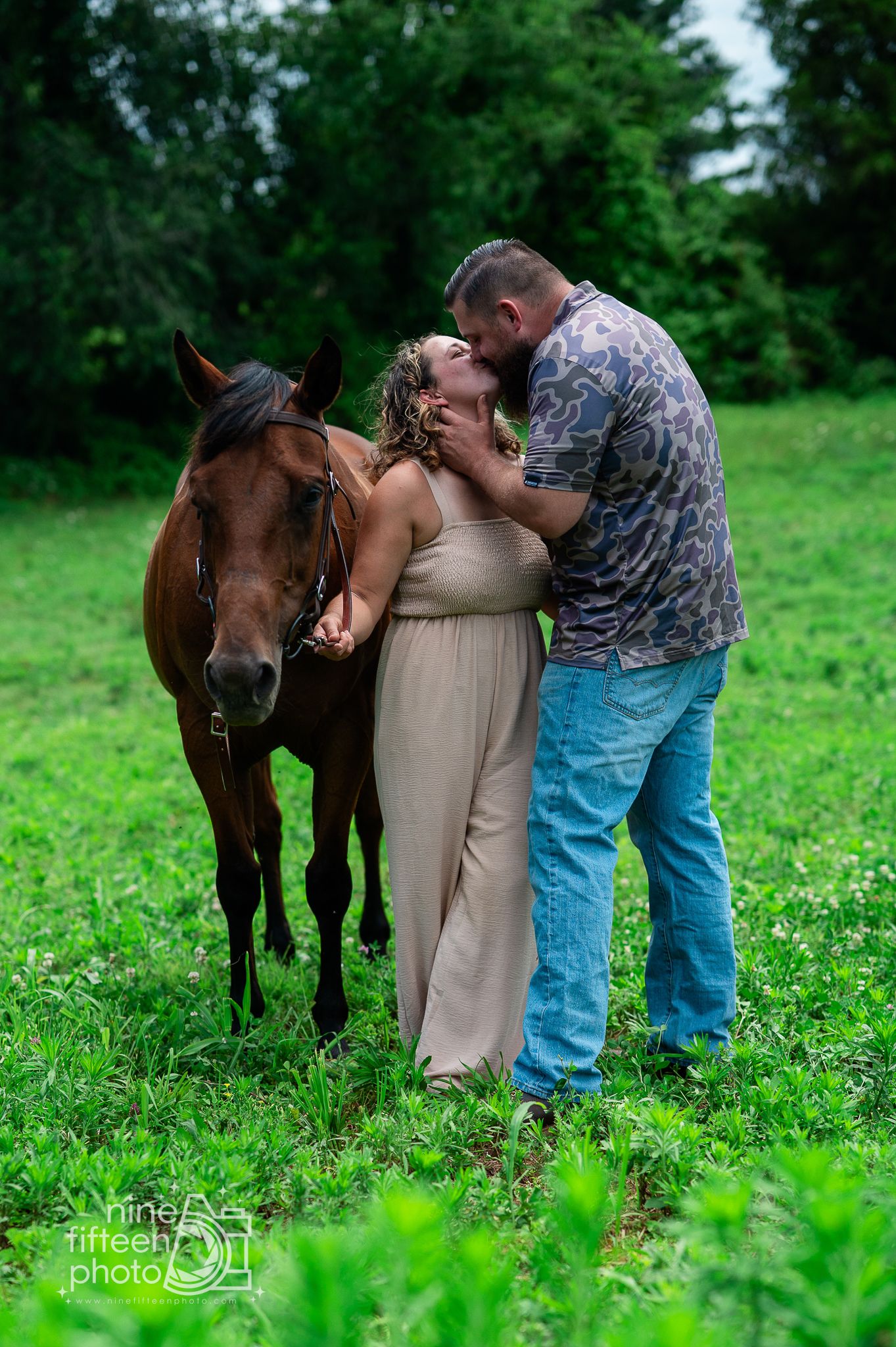 Couple sharing a romantic kiss beside a brown horse in a lush green field, captured by Nine Fifteen Photo.