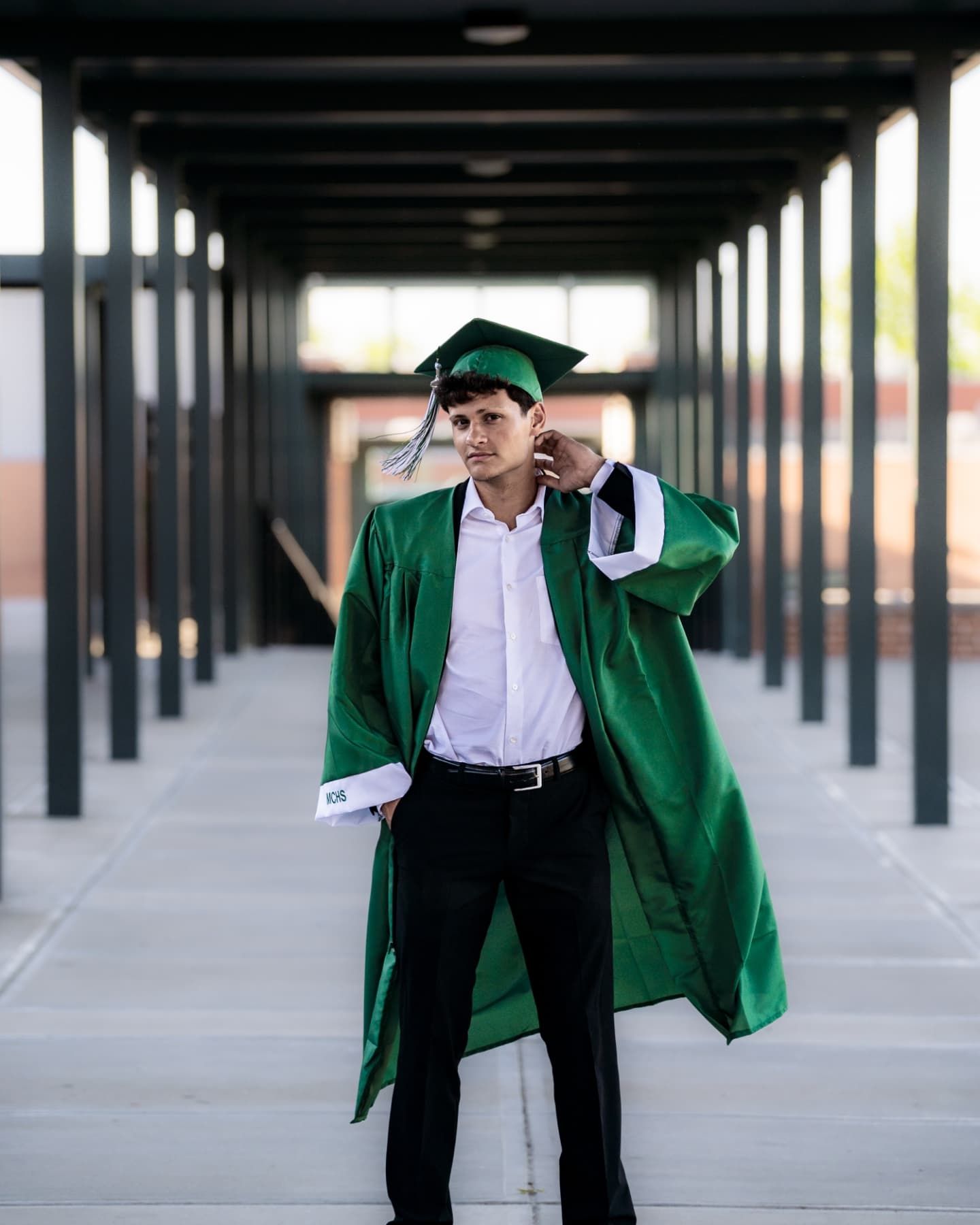 Male graduate in green cap and gown walks confidently through a covered outdoor corridor.