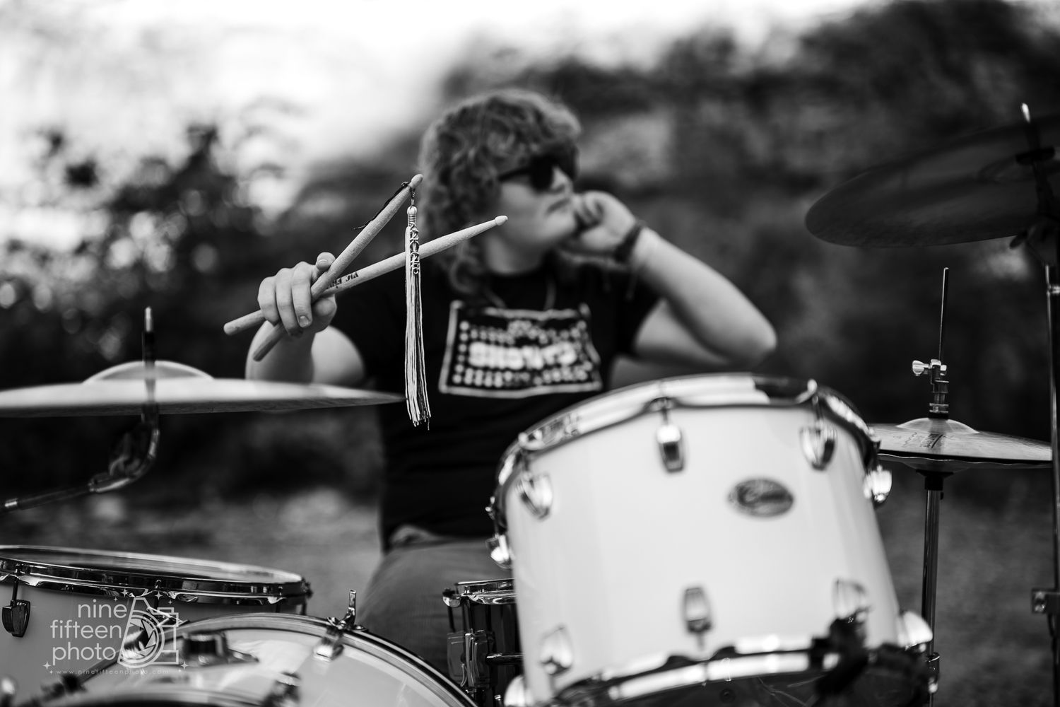 Black and white portrait of young drummer playing drums outdoors with sunglasses and drumsticks.