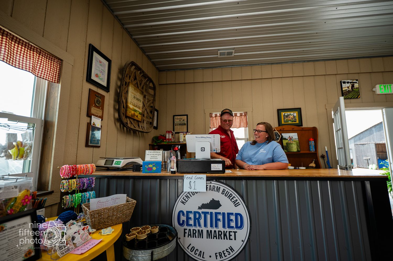 Two staff members stand behind counter at a Certified Farm Market with rustic wood-paneled interior.