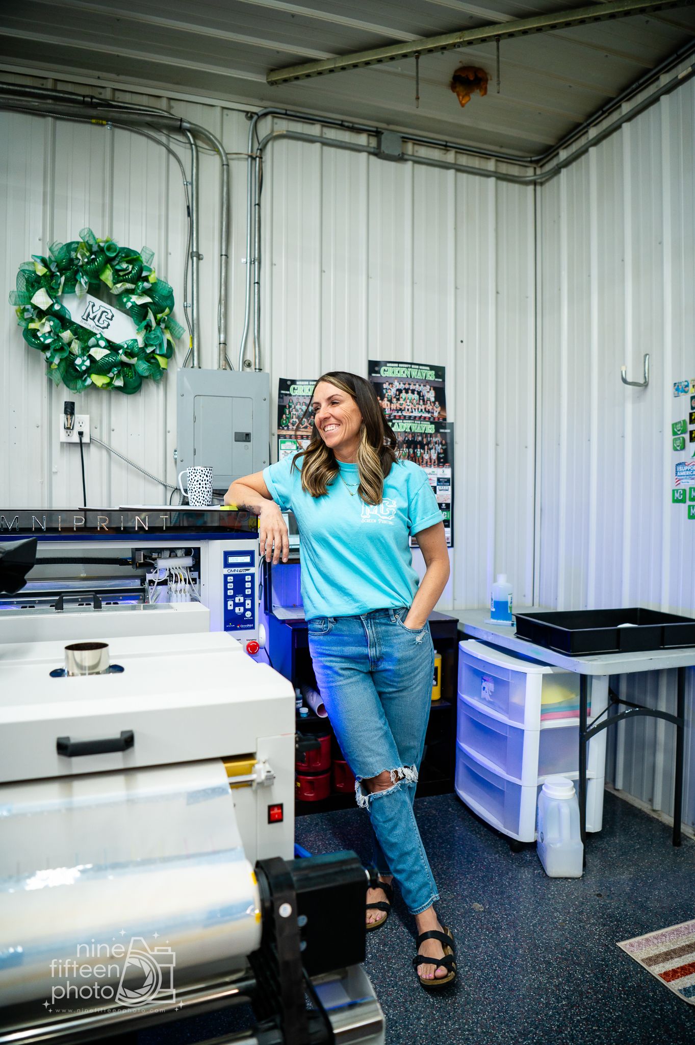Woman in teal shirt and jeans posing in a custom apparel printing shop.