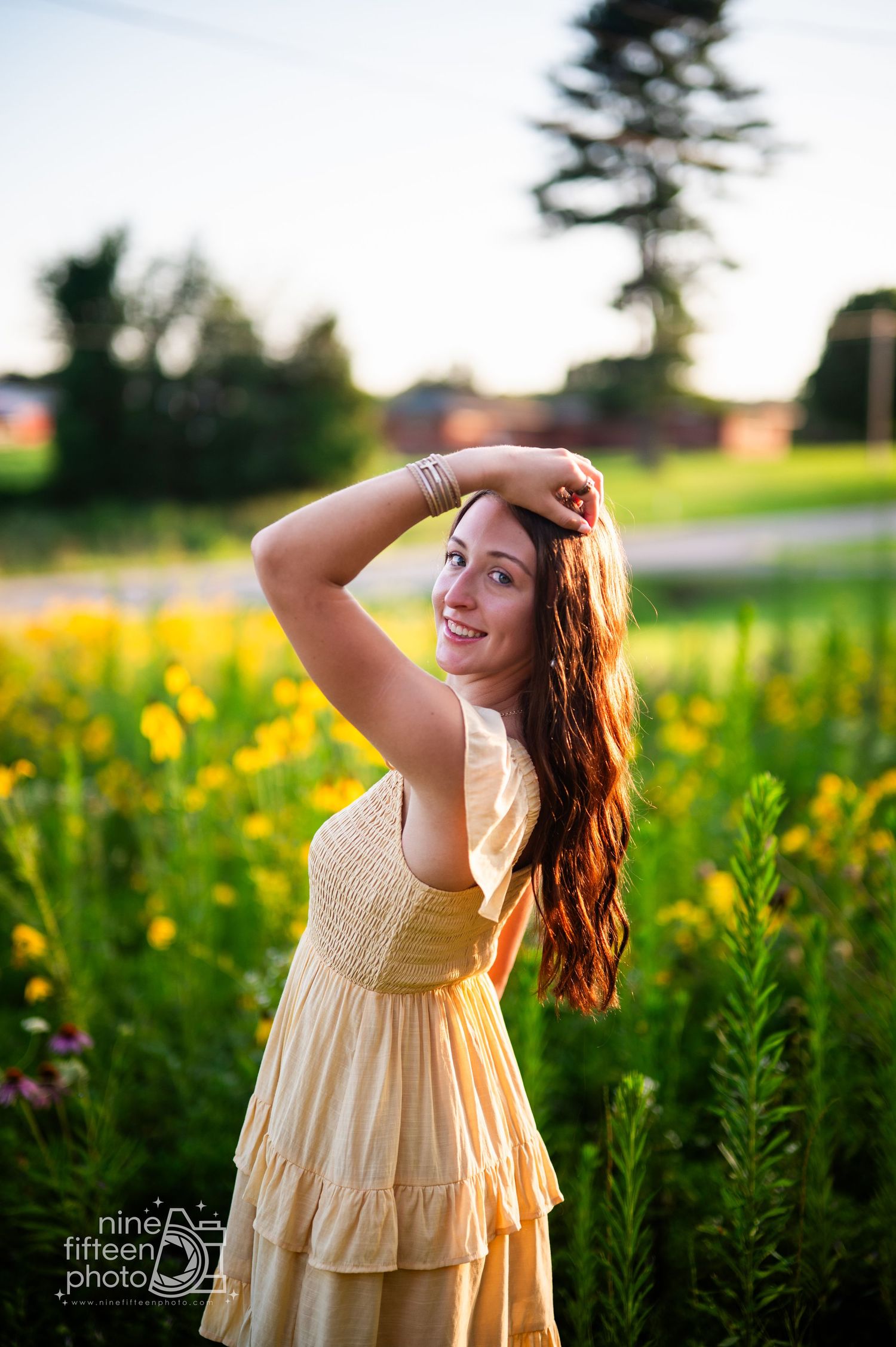 Young woman in yellow dress posing in a sunlit wildflower field, smiling with hand in hair.