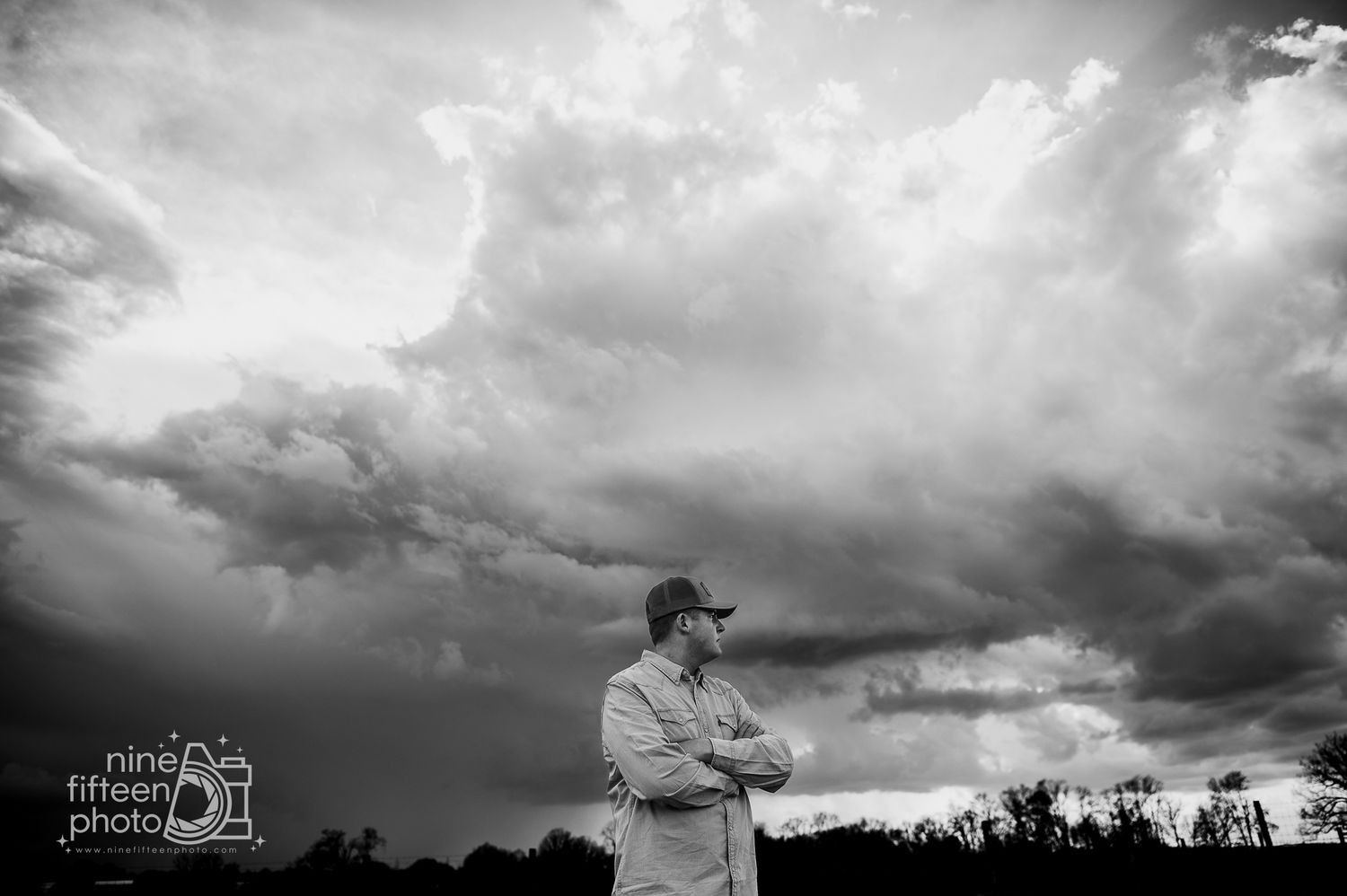 Black and white photo of a man with crossed arms standing under dramatic storm clouds in an open landscape.