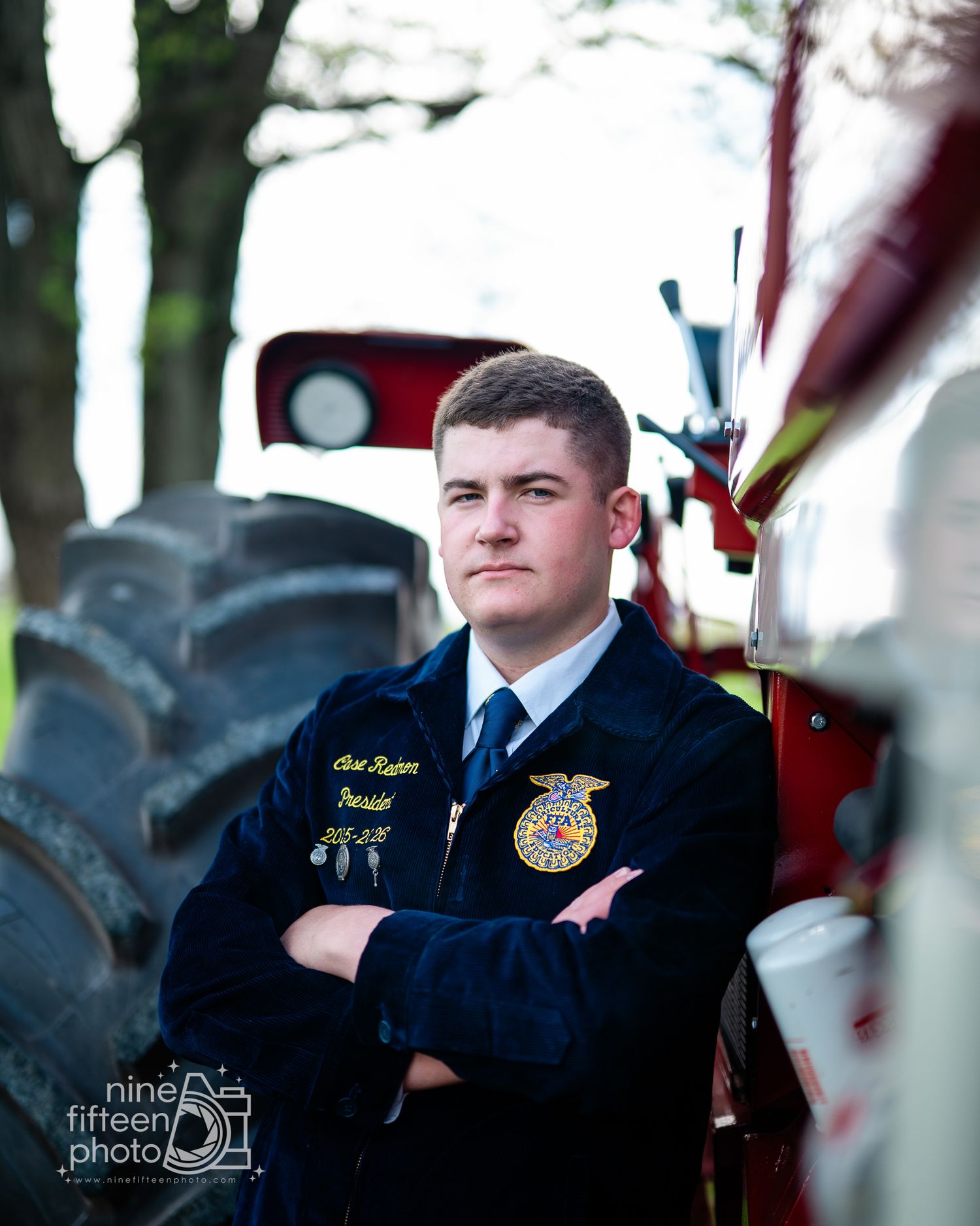 Young man in FFA jacket stands confidently with arms crossed beside a red tractor outdoors.