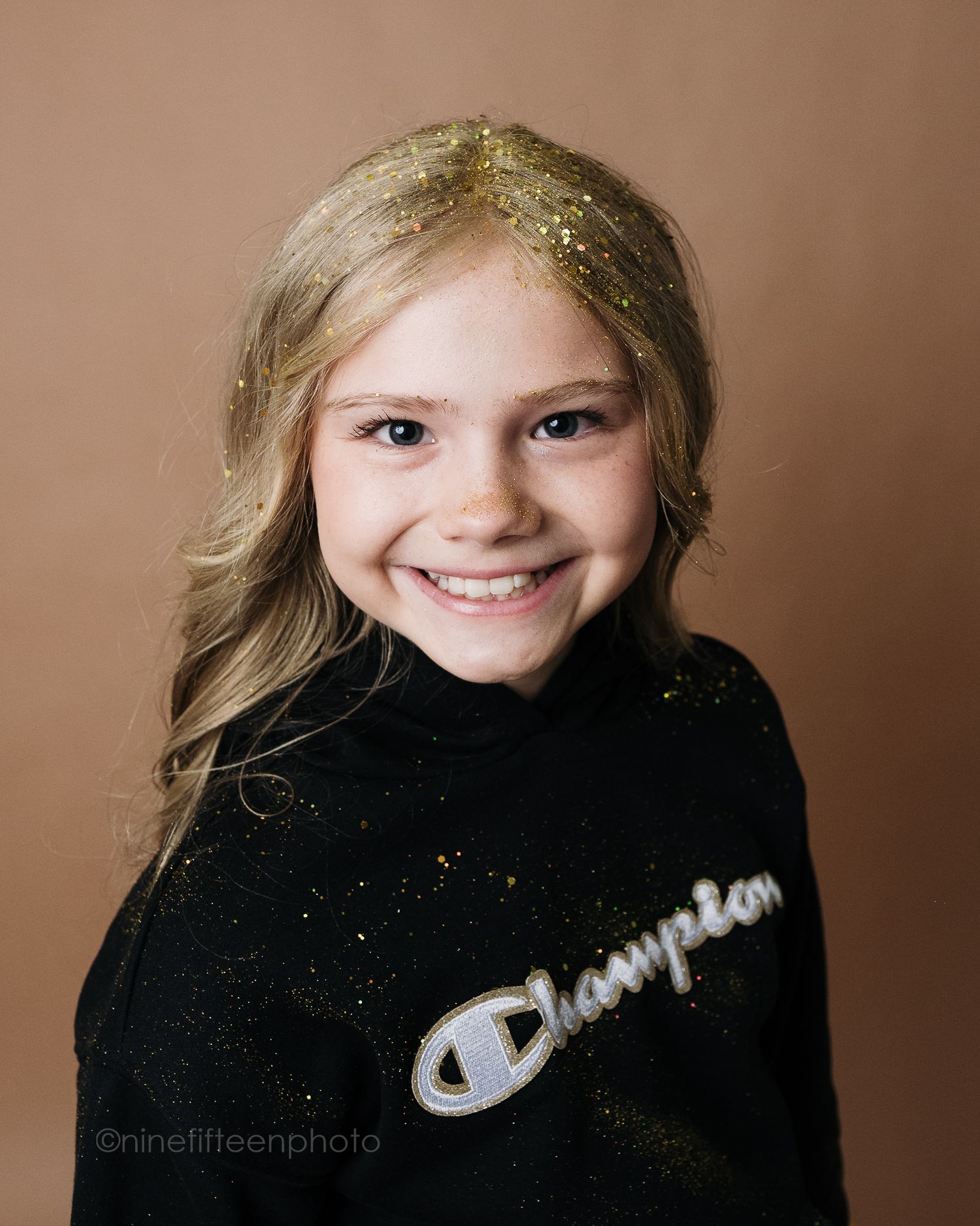 Smiling young girl with glitter in blonde hair wearing a black Champion sweatshirt against a tan background.