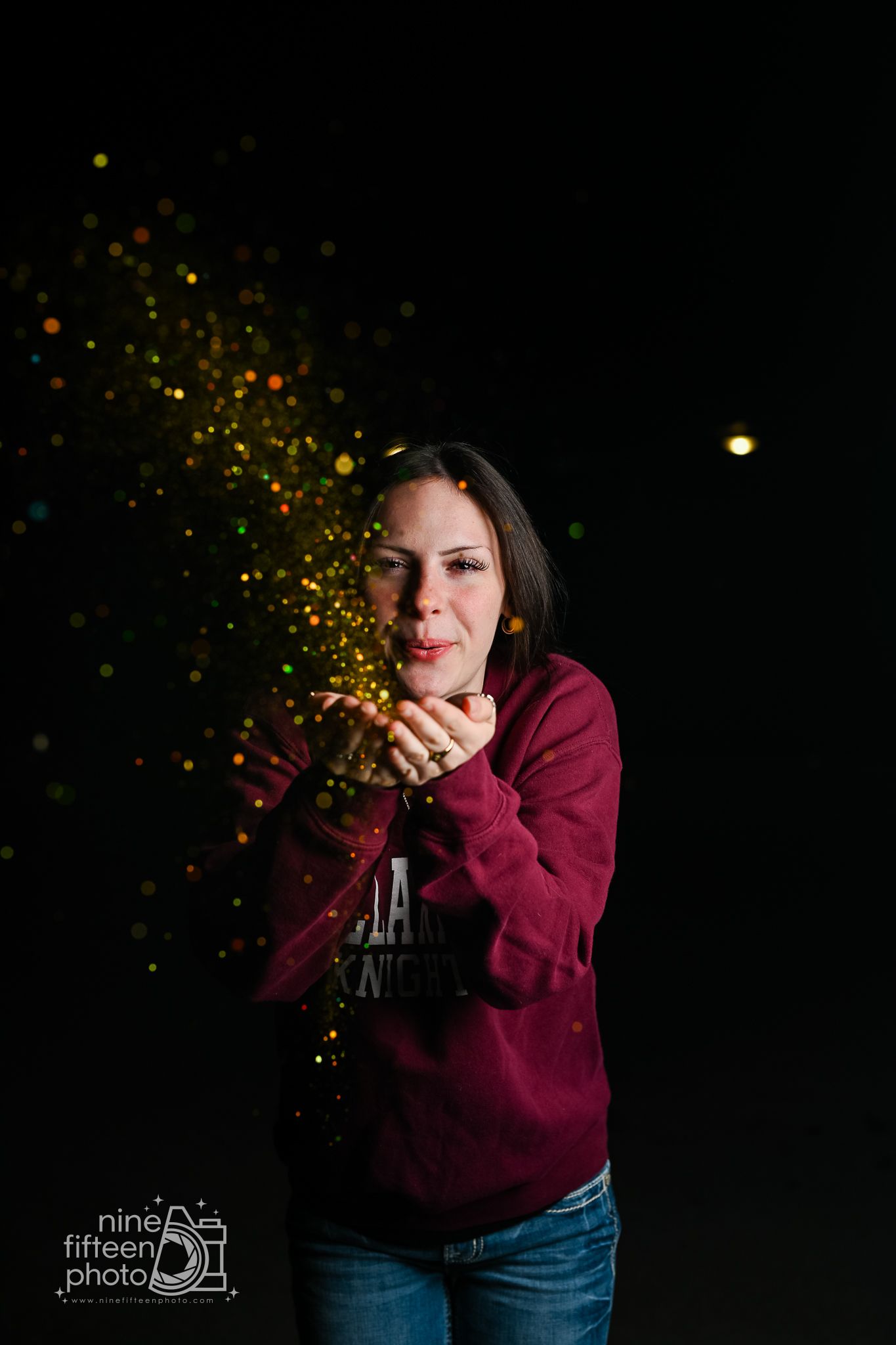 Woman in maroon sweatshirt blowing colorful glitter against dark background in studio portrait photo.