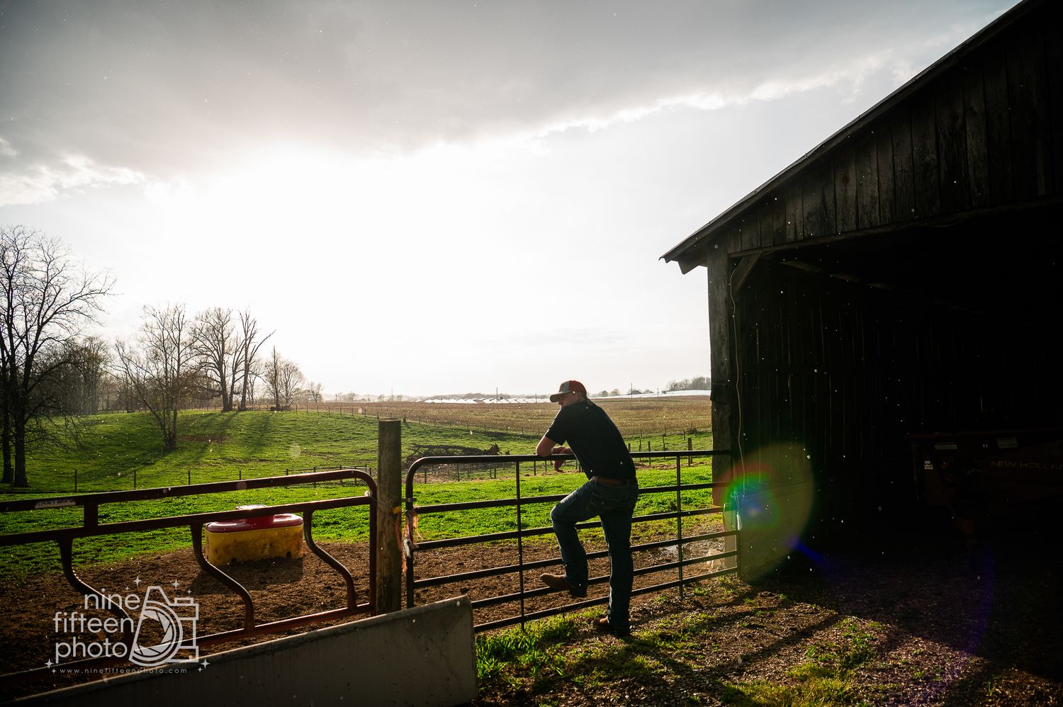 Person climbing a metal farm gate beside a rustic barn with green fields and dramatic cloudy sky in background.
