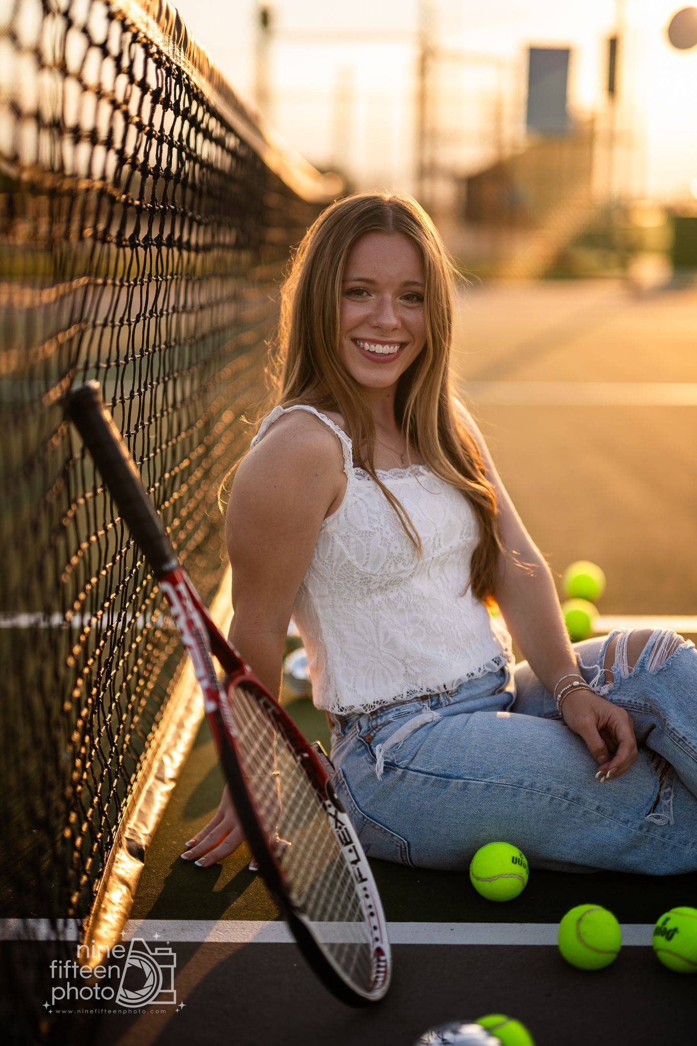 Smiling teenage girl sitting by tennis net with racket and yellow balls at golden sunset.