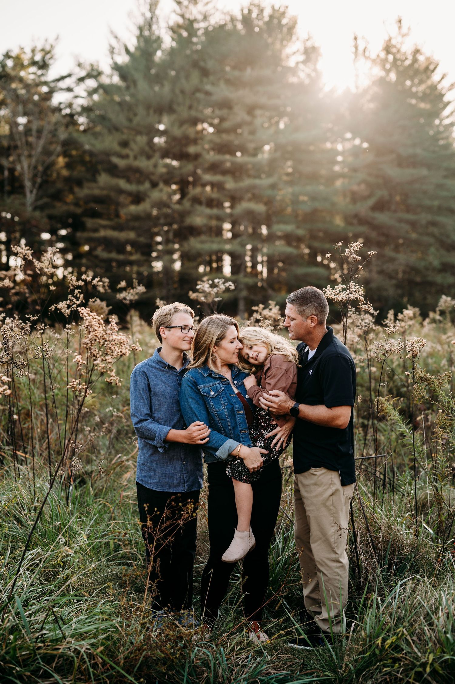 Family of four shares tender moment in sunlit meadow surrounded by tall grass and pine trees.