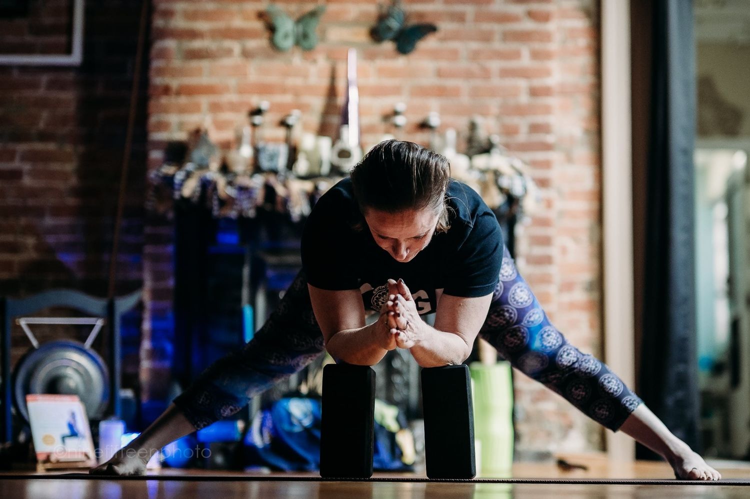 Woman performing a deep yoga lunge pose on a mat in a rustic brick-walled studio.
