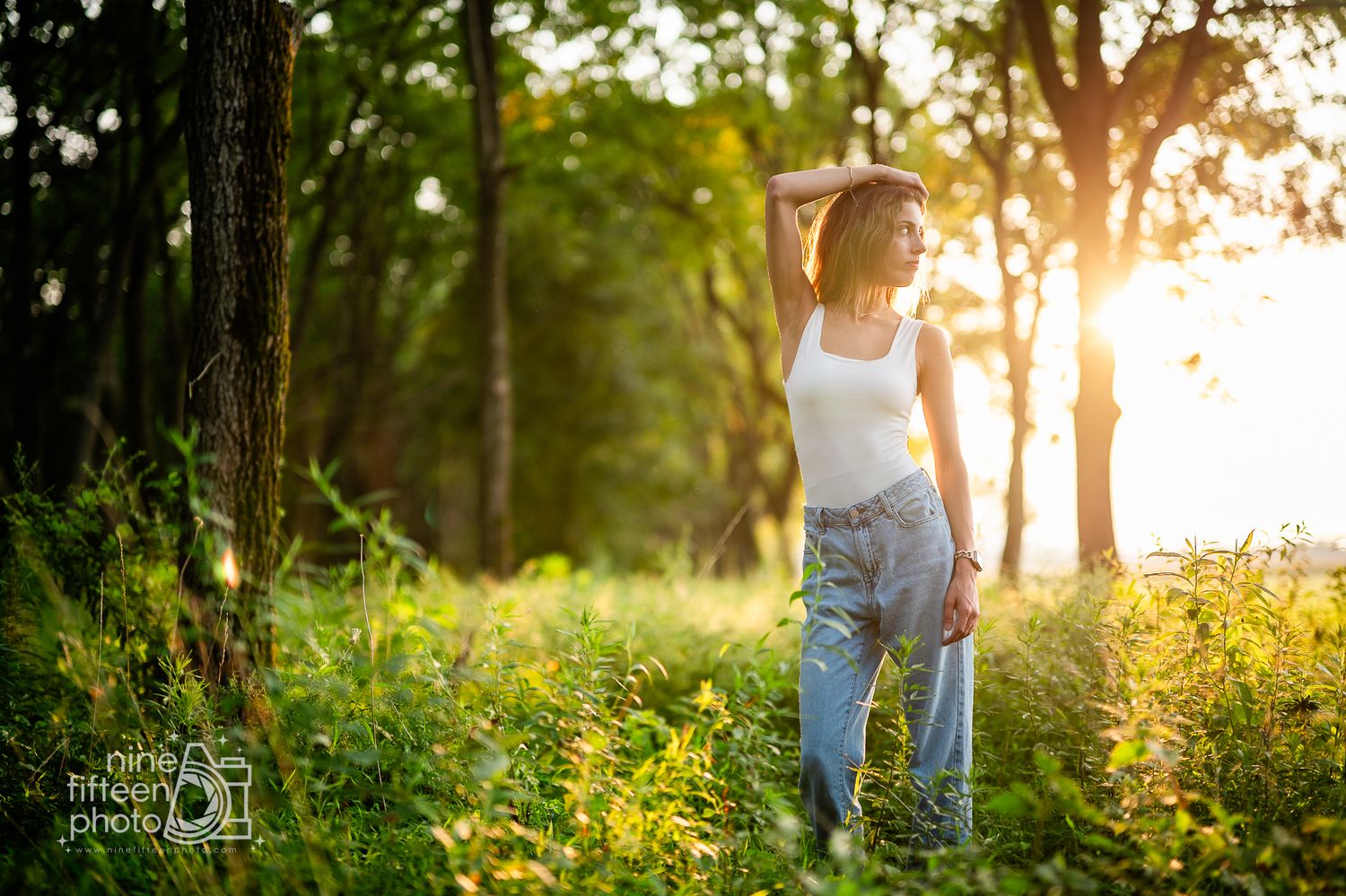 Young woman in white top and jeans poses in sunlit forest clearing surrounded by wildflowers and golden light.
