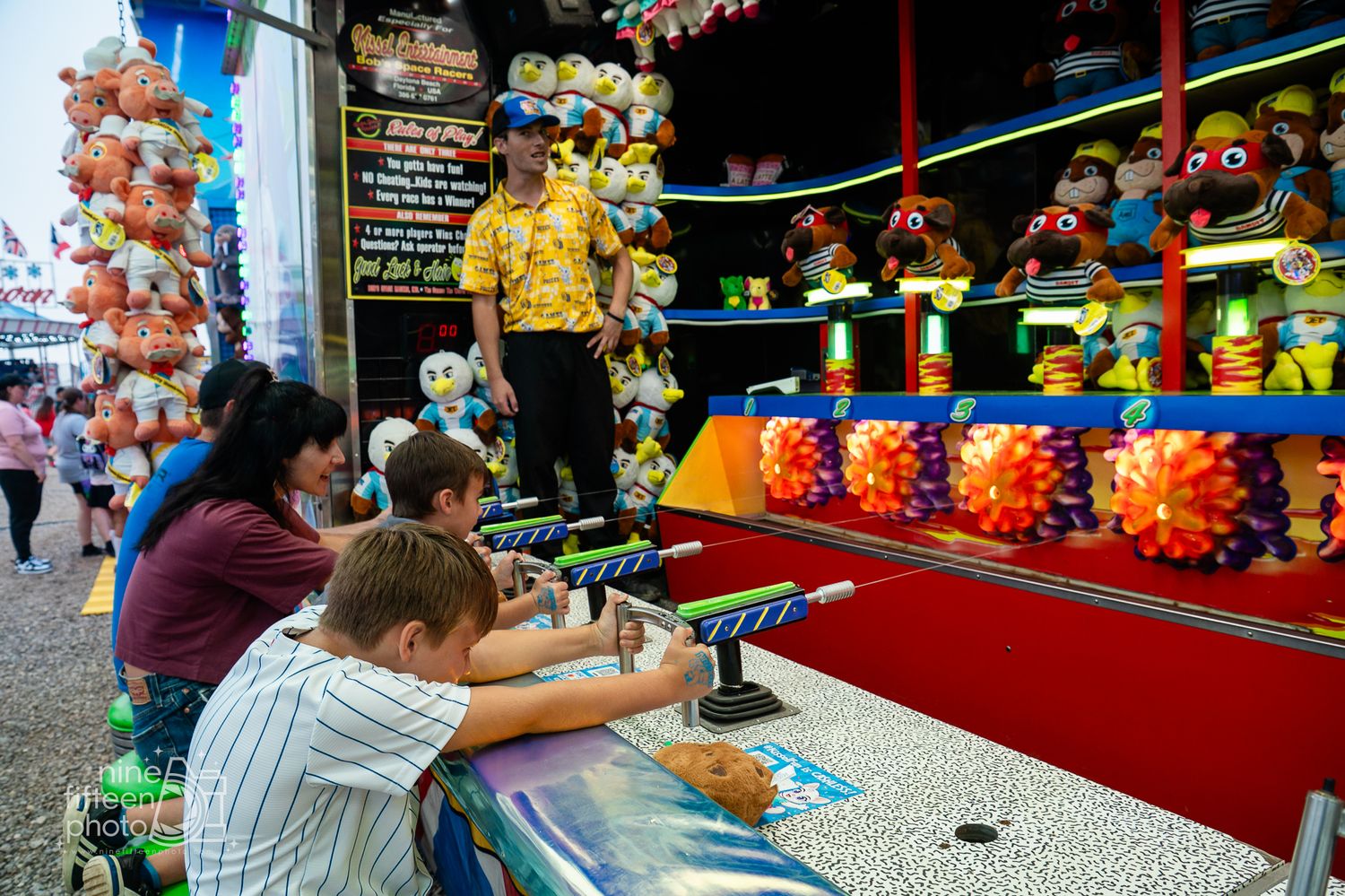 Kids playing a shooting gallery game at a colorful carnival midway with stuffed animal prizes on display.