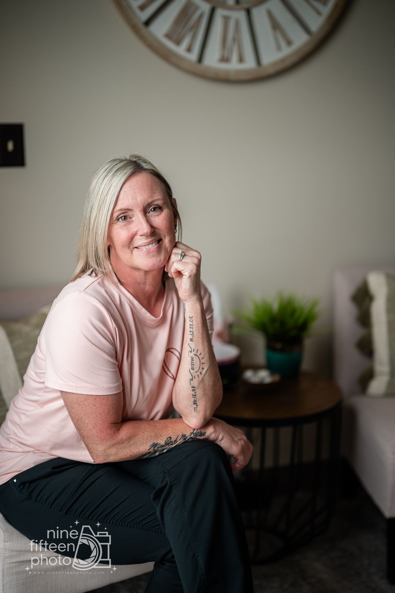 Smiling blonde woman with tattoos on her arm poses indoors near a decorative clock and round side table.