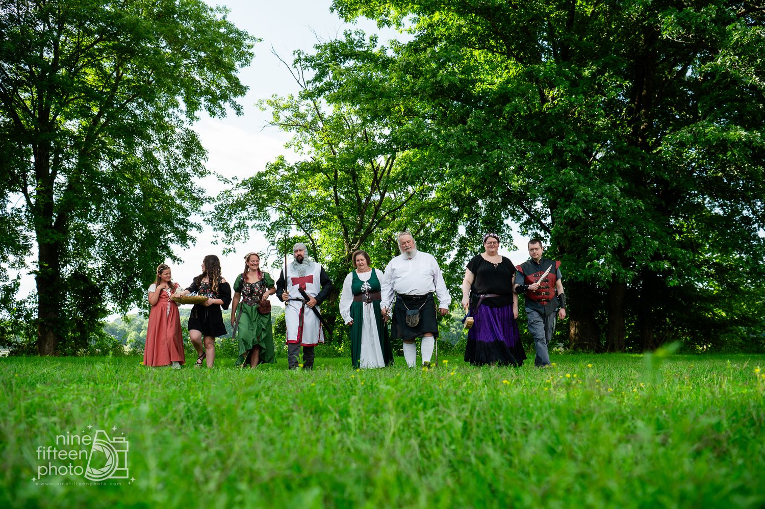 Group of people in medieval Renaissance costumes posing outdoors on green grass with lush trees in background.