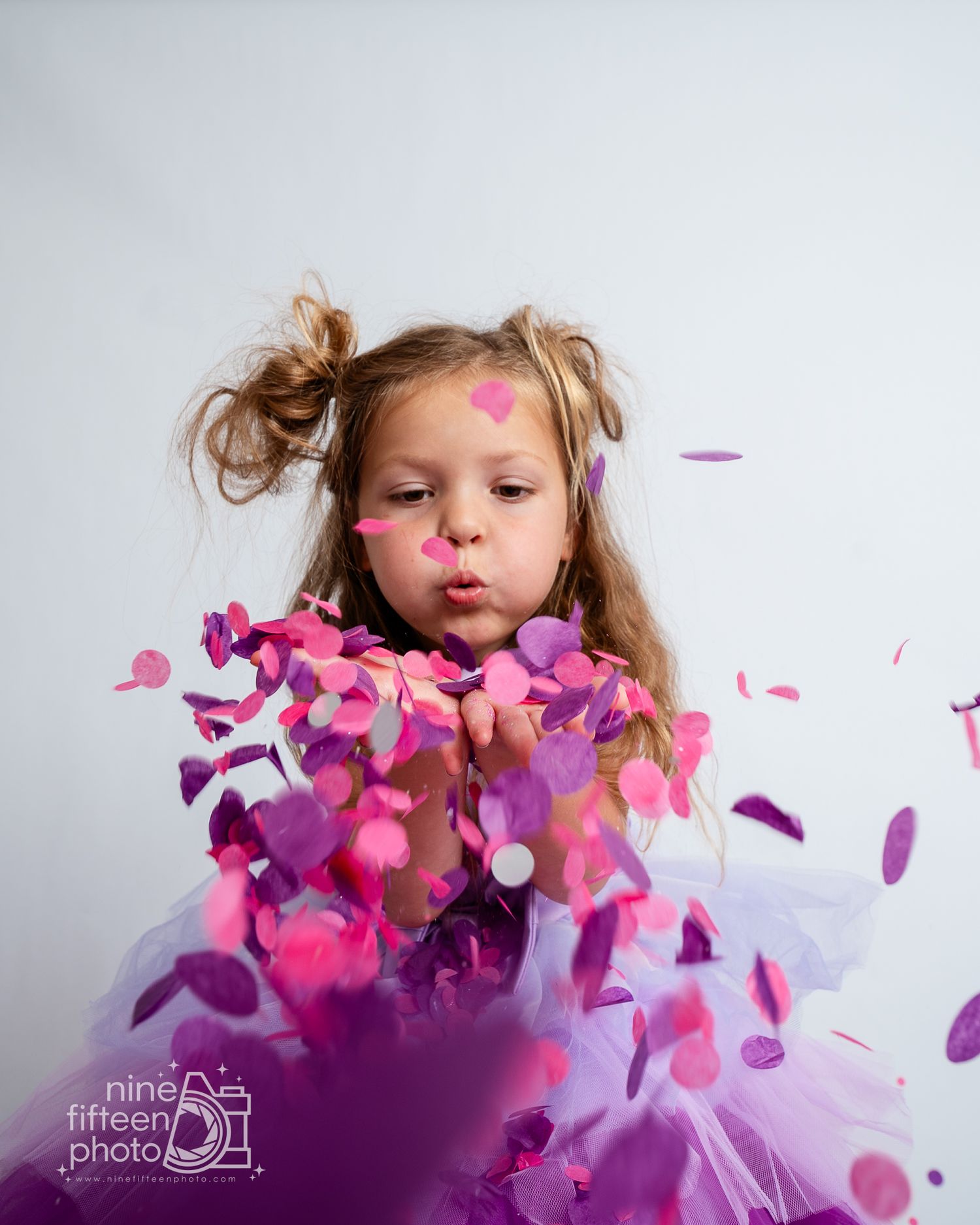 Young girl in purple tutu blowing pink and purple confetti, captured in a whimsical studio portrait.