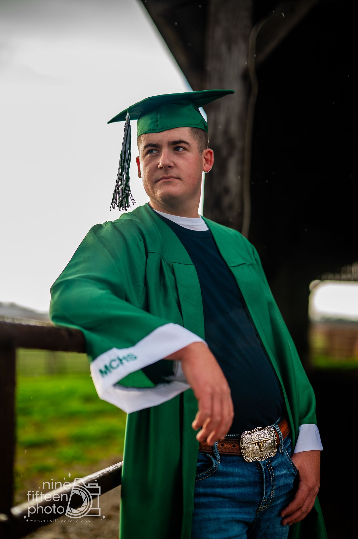 Young man in green graduation cap and gown with MCHS insignia posing outdoors for senior portrait photo.