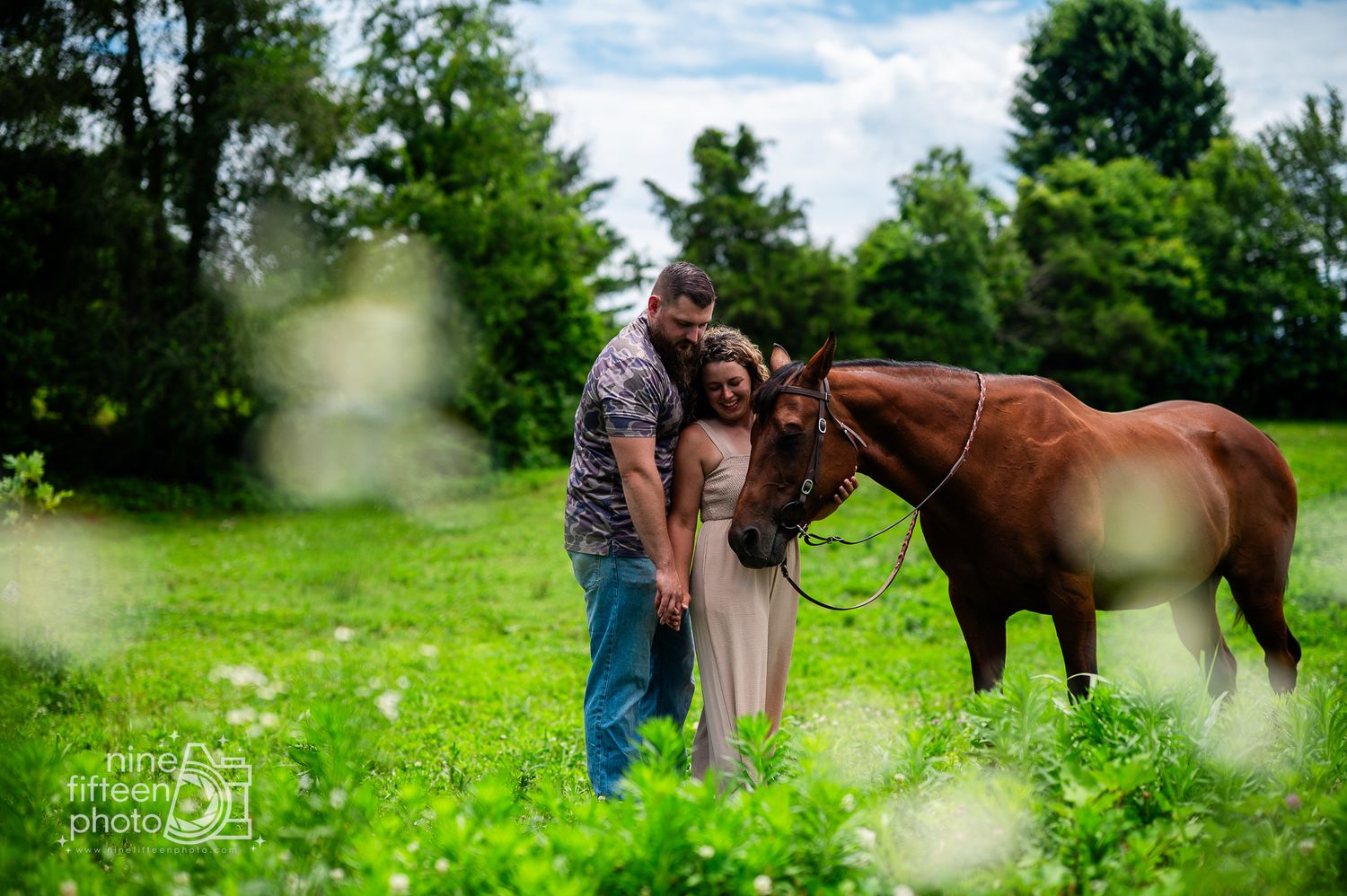 Couple standing together with a brown horse in a lush green field during an outdoor engagement photo session.