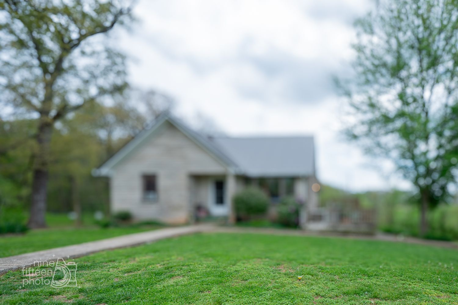 Blurred rustic white farmhouse with gray roof surrounded by lush green lawn and trees on a cloudy day.