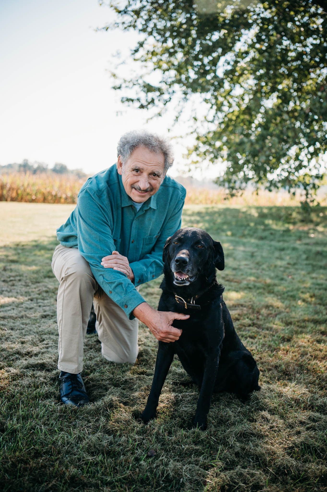 Grandfather kneeling beside black family dog Boomer during outdoor fall photo session in Meade County Kentucky.