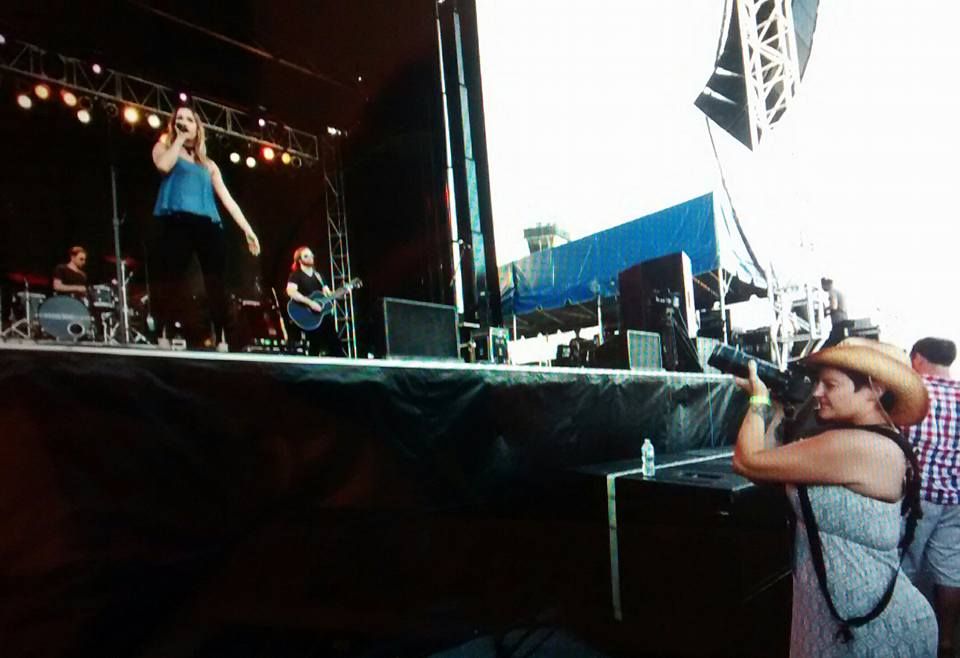Female photographer capturing concert performance on outdoor stage at music festival.