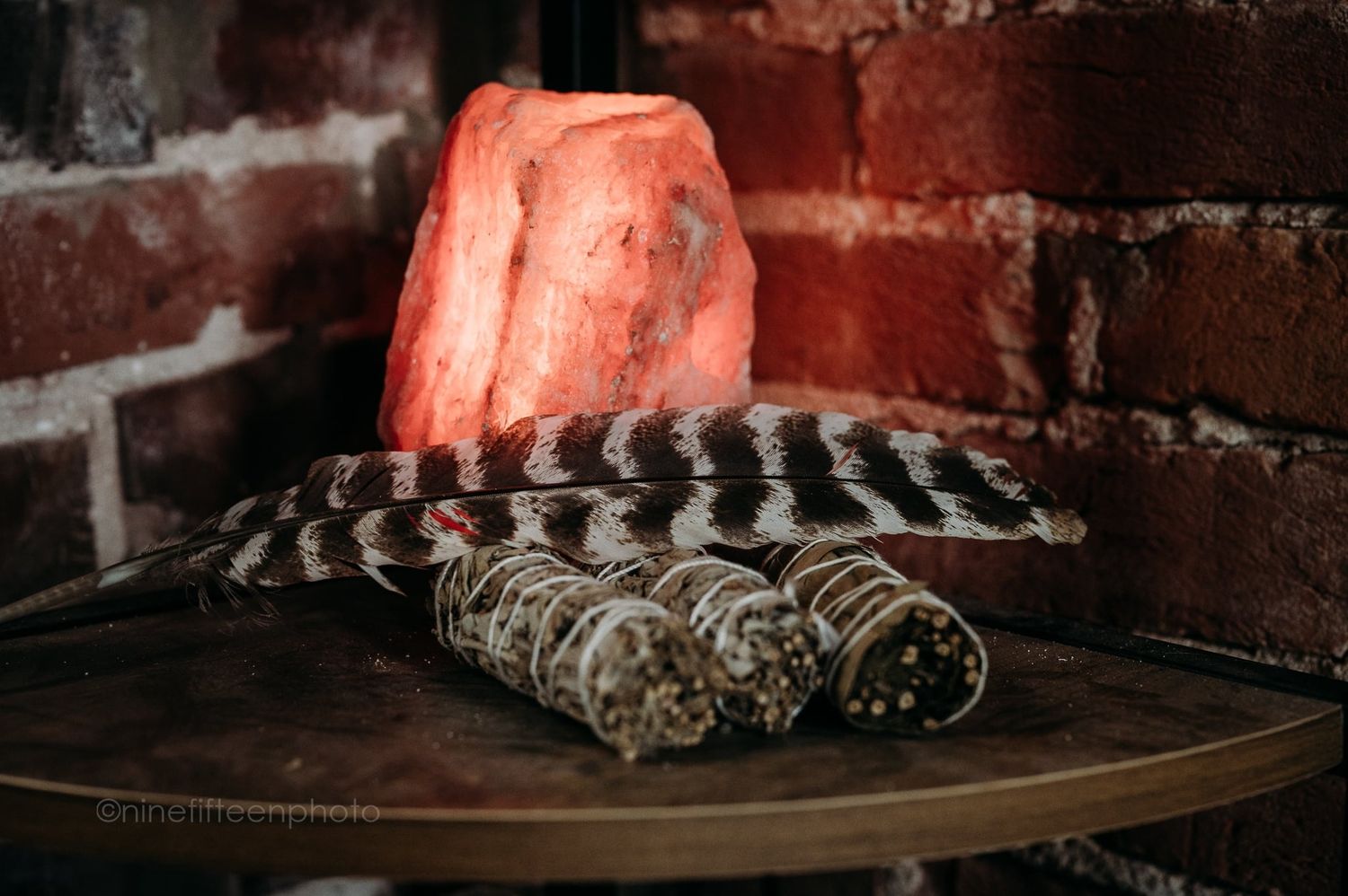 Himalayan salt lamp glows pink beside feathers and sage bundles on wooden table against brick wall.
