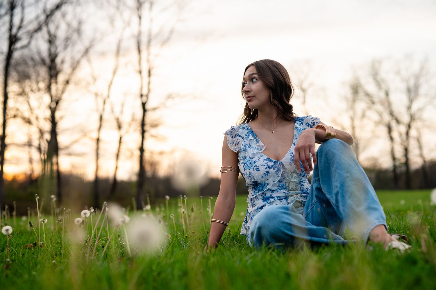 Young woman in blue floral top and jeans sitting on lush green grass at sunset in a peaceful park.