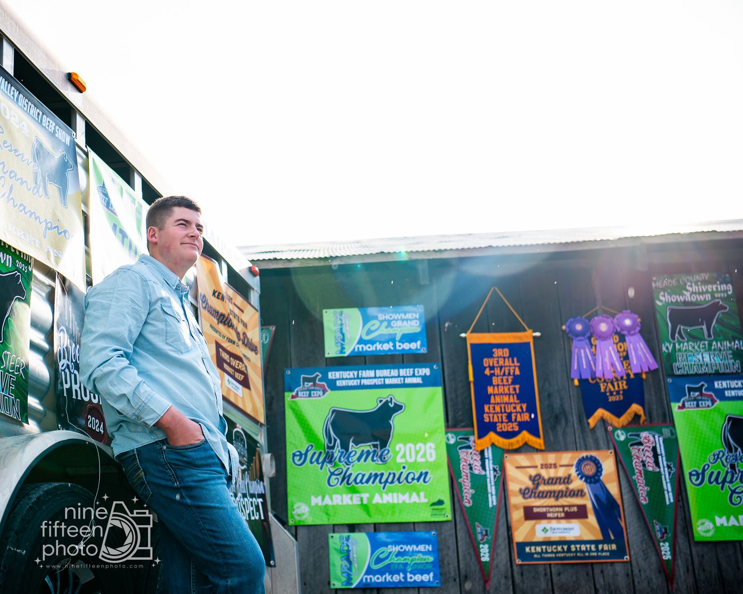 Young man in light blue jacket stands proudly before colorful fair banners and Champion Market Animal signs at an agricultural event.