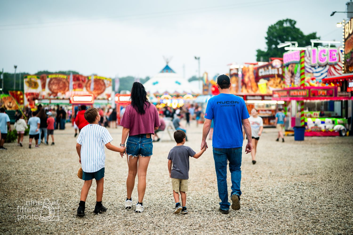 Family of four holding hands walking through a colorful carnival fairground with game booths and rides in the background.