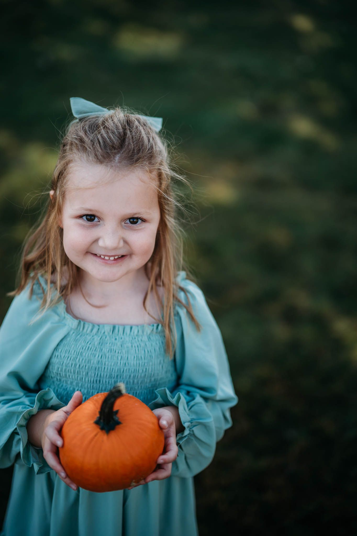 Little girl in blue dress holding a small orange pumpkin during autumn family photo session.