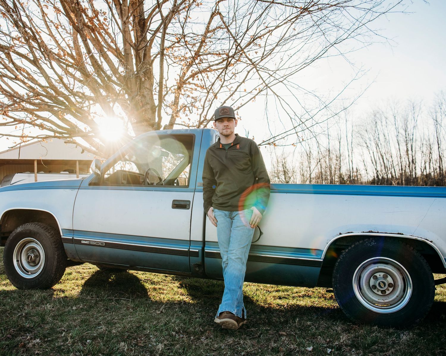 Senior leaning against vintage truck at golden sunset in open Kentucky field.