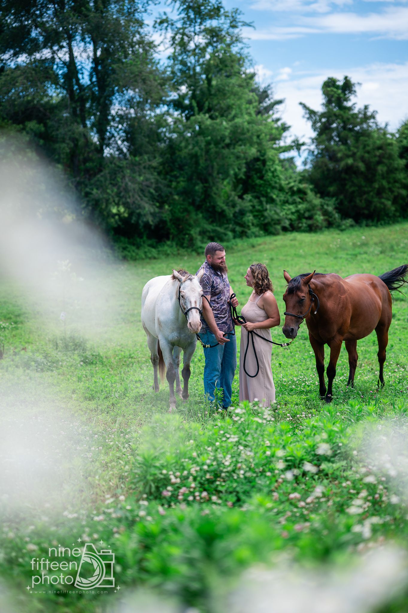 Couple standing together in a lush green field holding a white and brown horse, outdoor engagement photo session.