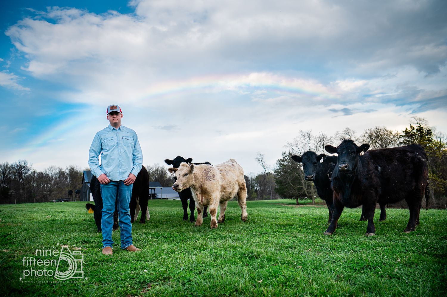 A farmer stands in a green pasture with black and white cattle under a partly cloudy sky.