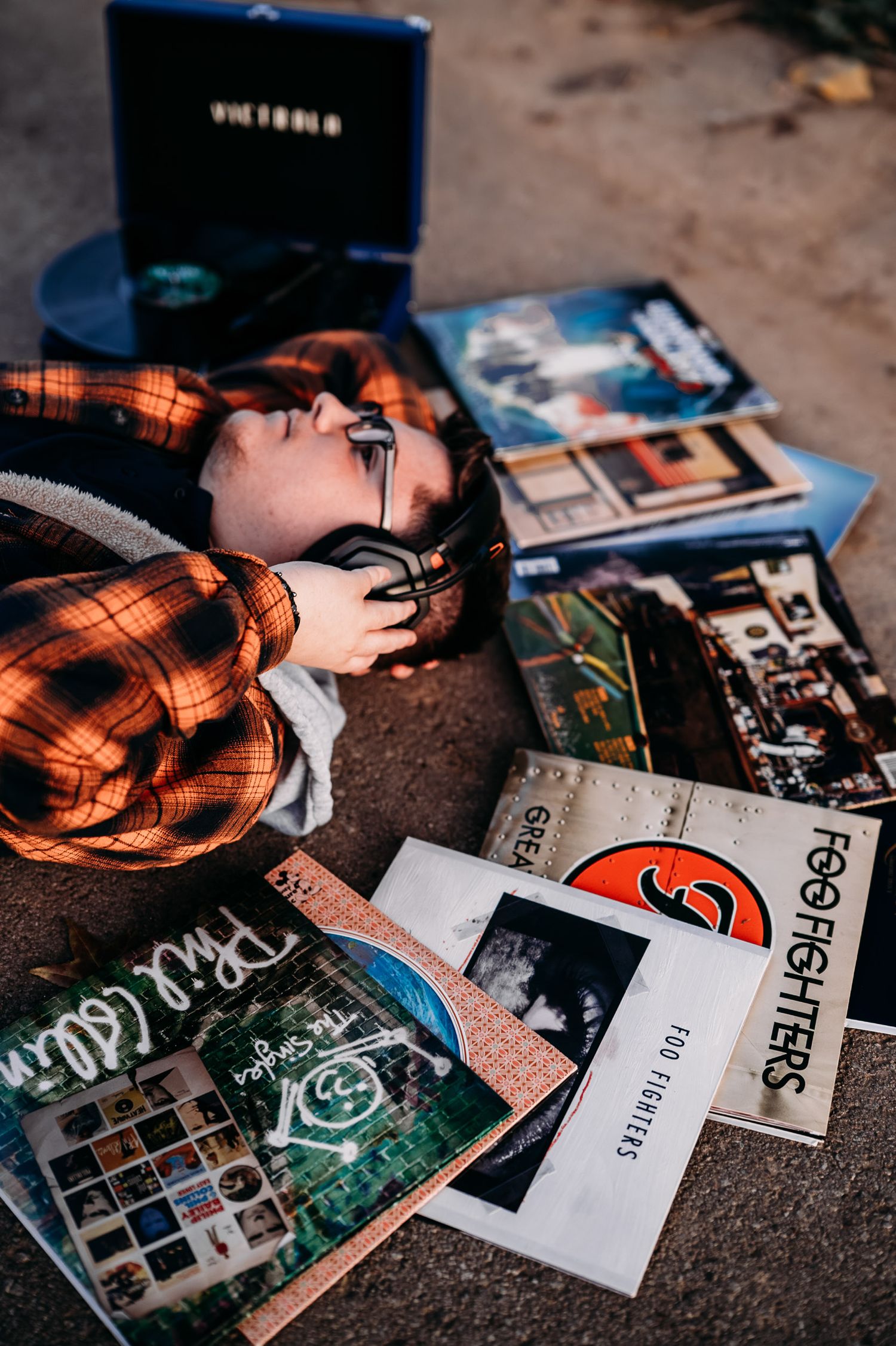 Person lying among vinyl records including Foo Fighters albums beside a blue portable turntable on pavement.