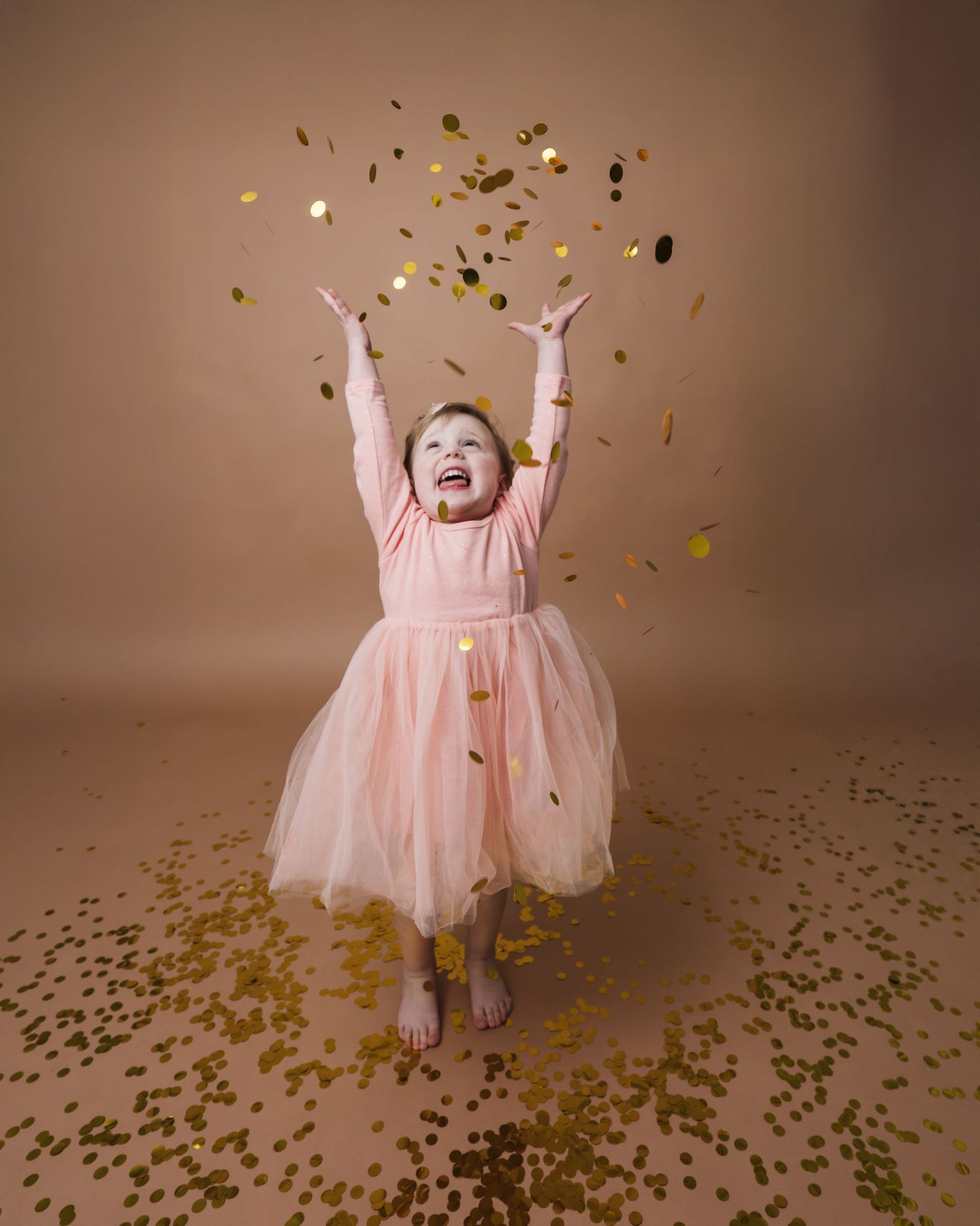 A joyful young girl in a pink tulle dress laughs as golden confetti rains down around her on a beige background.