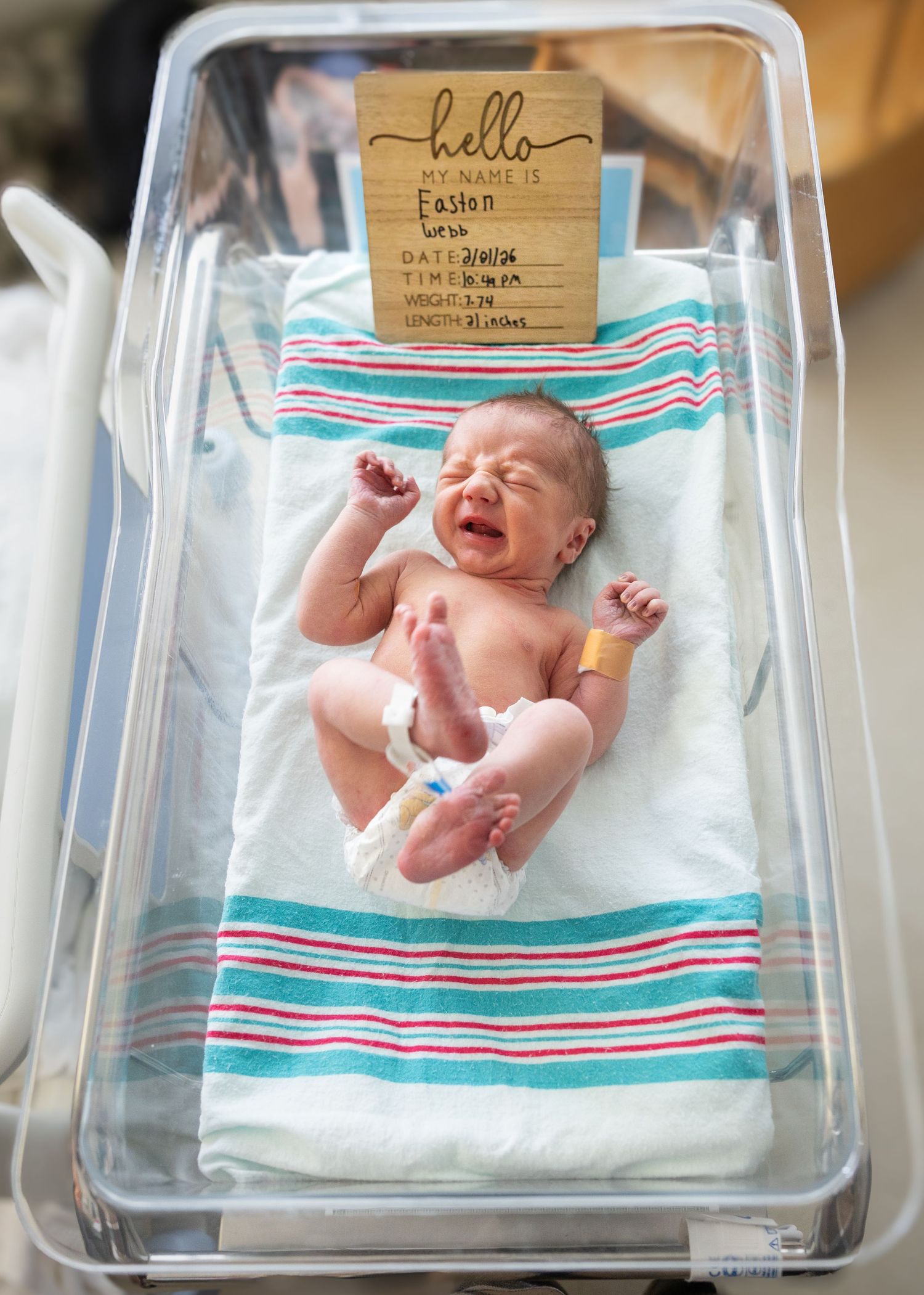 A newborn baby named Easton stretches in a hospital bassinet beside a personalized hello name sign.