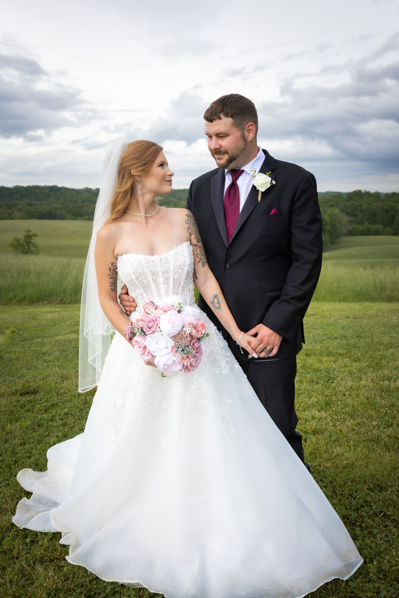 A bride and groom share a tender moment in a green field, bride holding a pink bouquet.