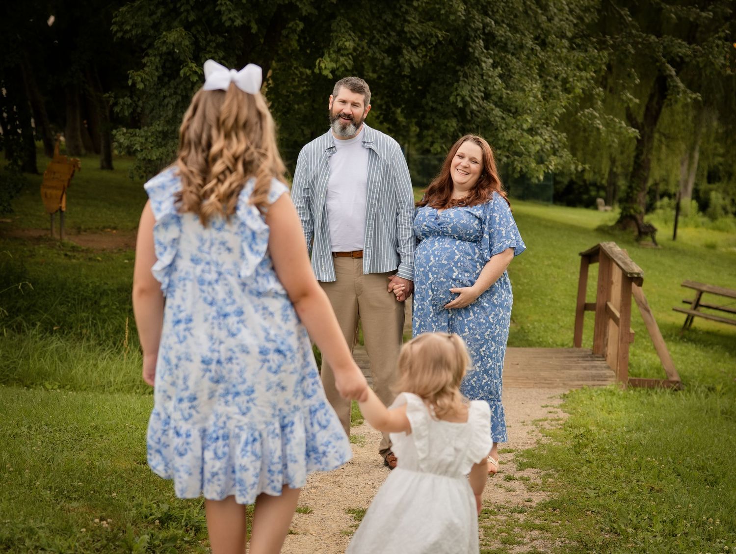 Pregnant woman and man hold hands with two young girls in blue dresses on a park path.