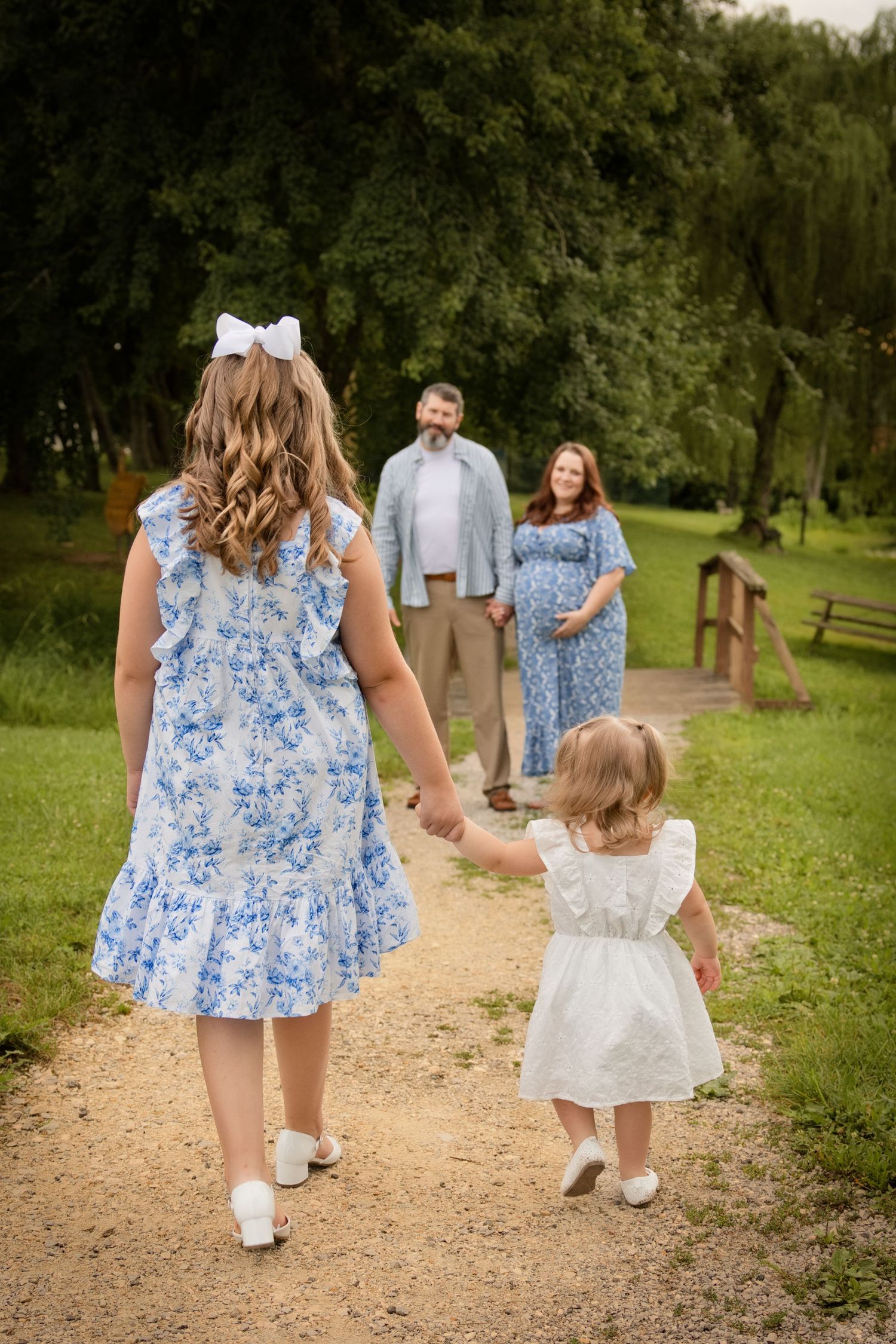 Two girls in blue and white dresses walk toward their pregnant mother and father on a park path.