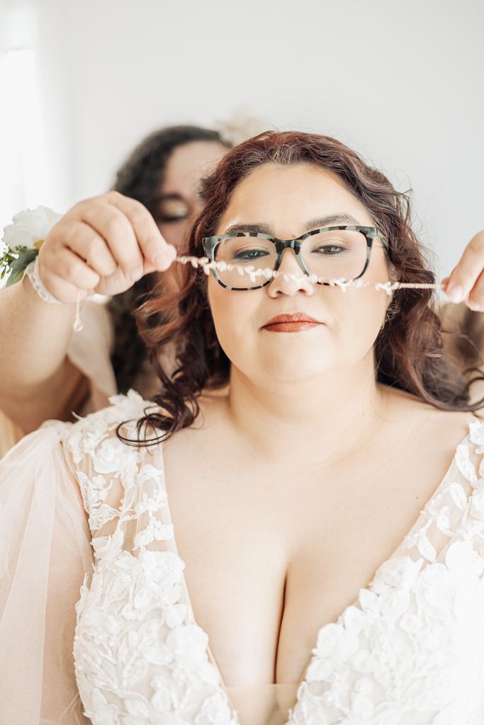 Plus-size bride in lace gown wearing glasses as bridesmaid places floral hair accessory.