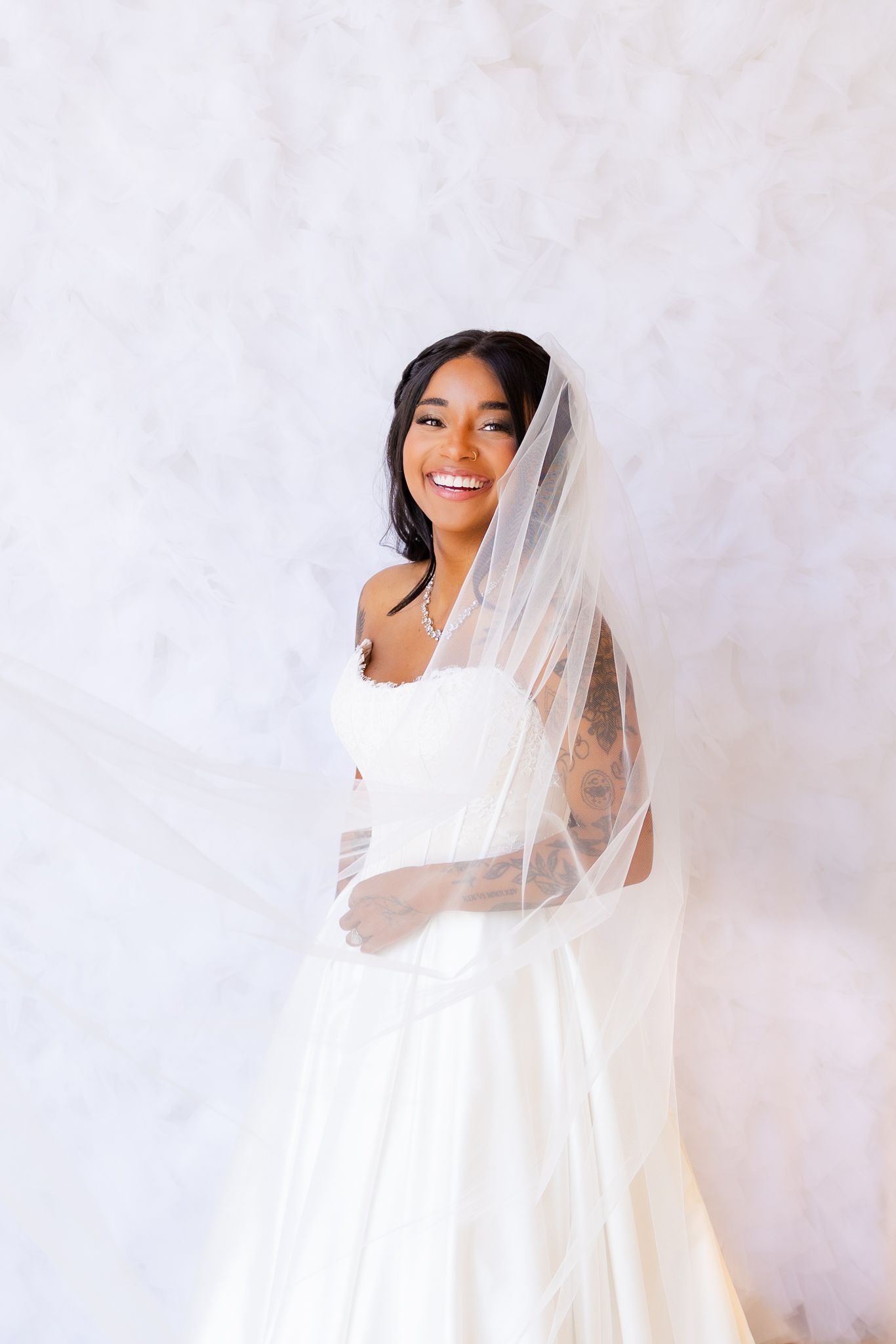 Smiling bride in white strapless gown and flowing veil poses against marble background.