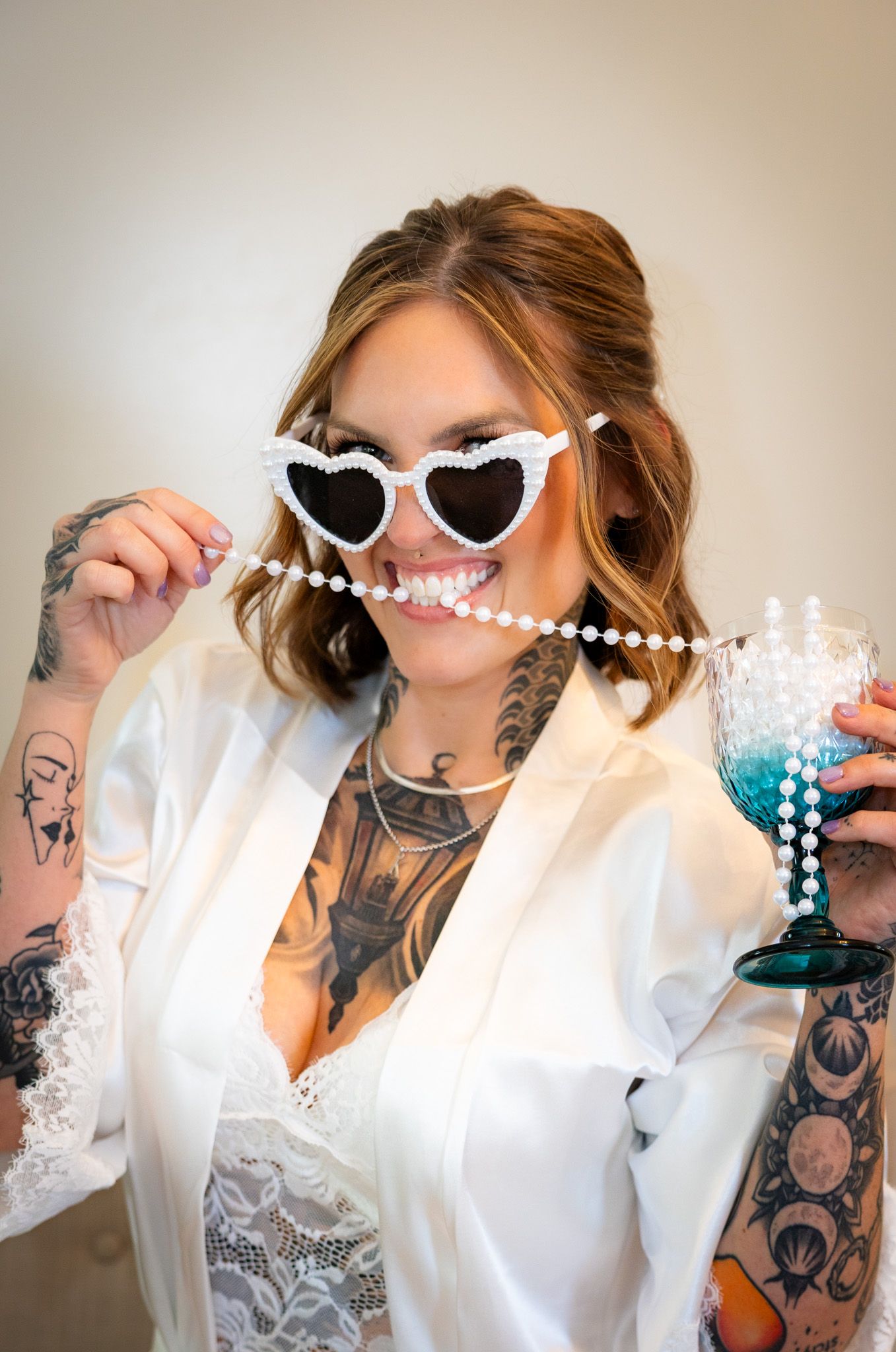 Tattooed bride in white robe poses playfully with pearl heart sunglasses and colorful cocktail.