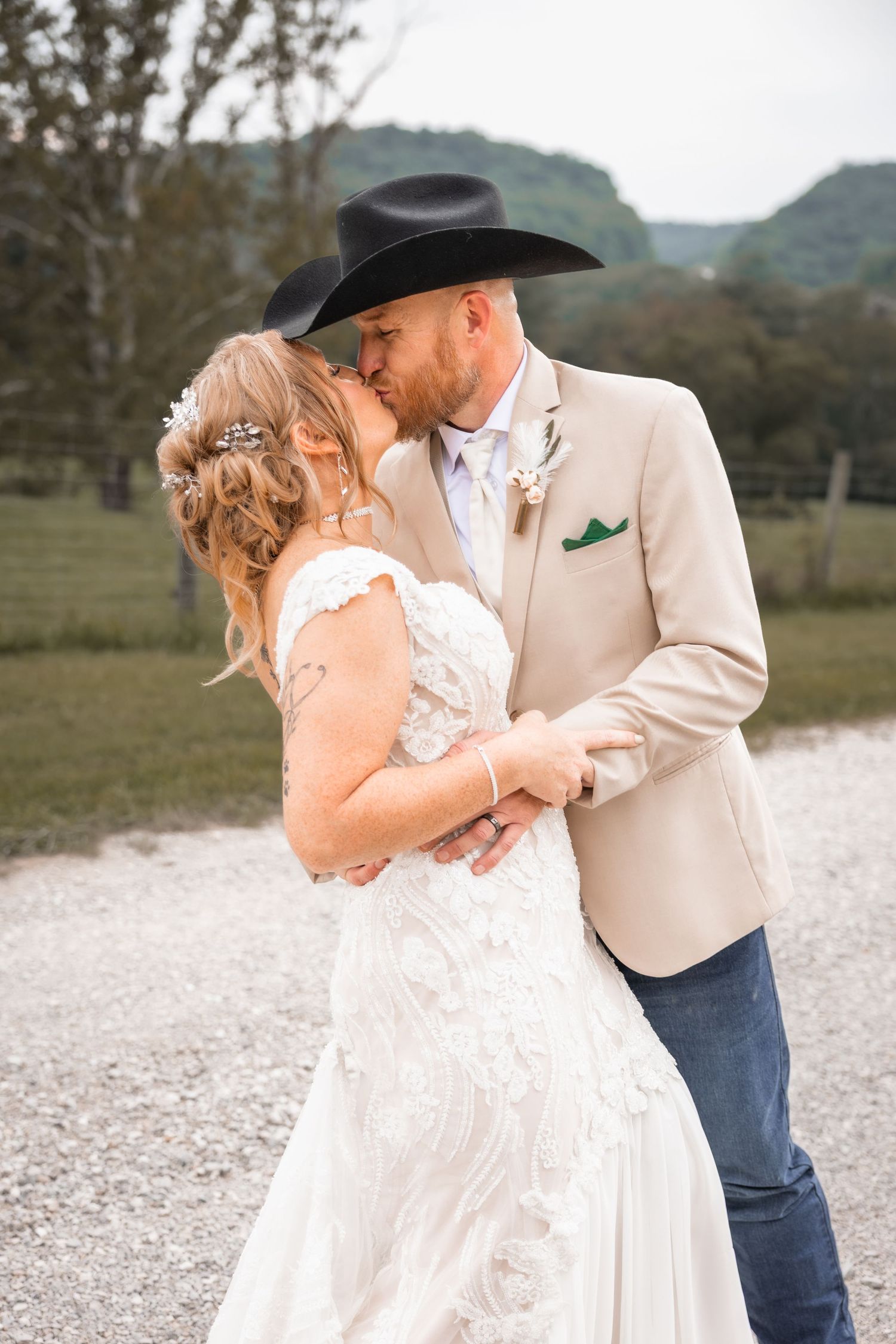 A bride and groom share a kiss outdoors, the groom wearing a black cowboy hat and tan blazer with a green pocket square.