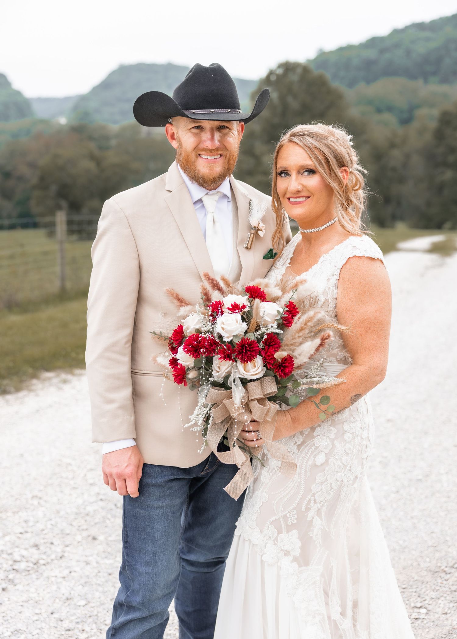 Bride in lace gown holds red and white bouquet beside groom in tan suit and black cowboy hat on rustic gravel path.