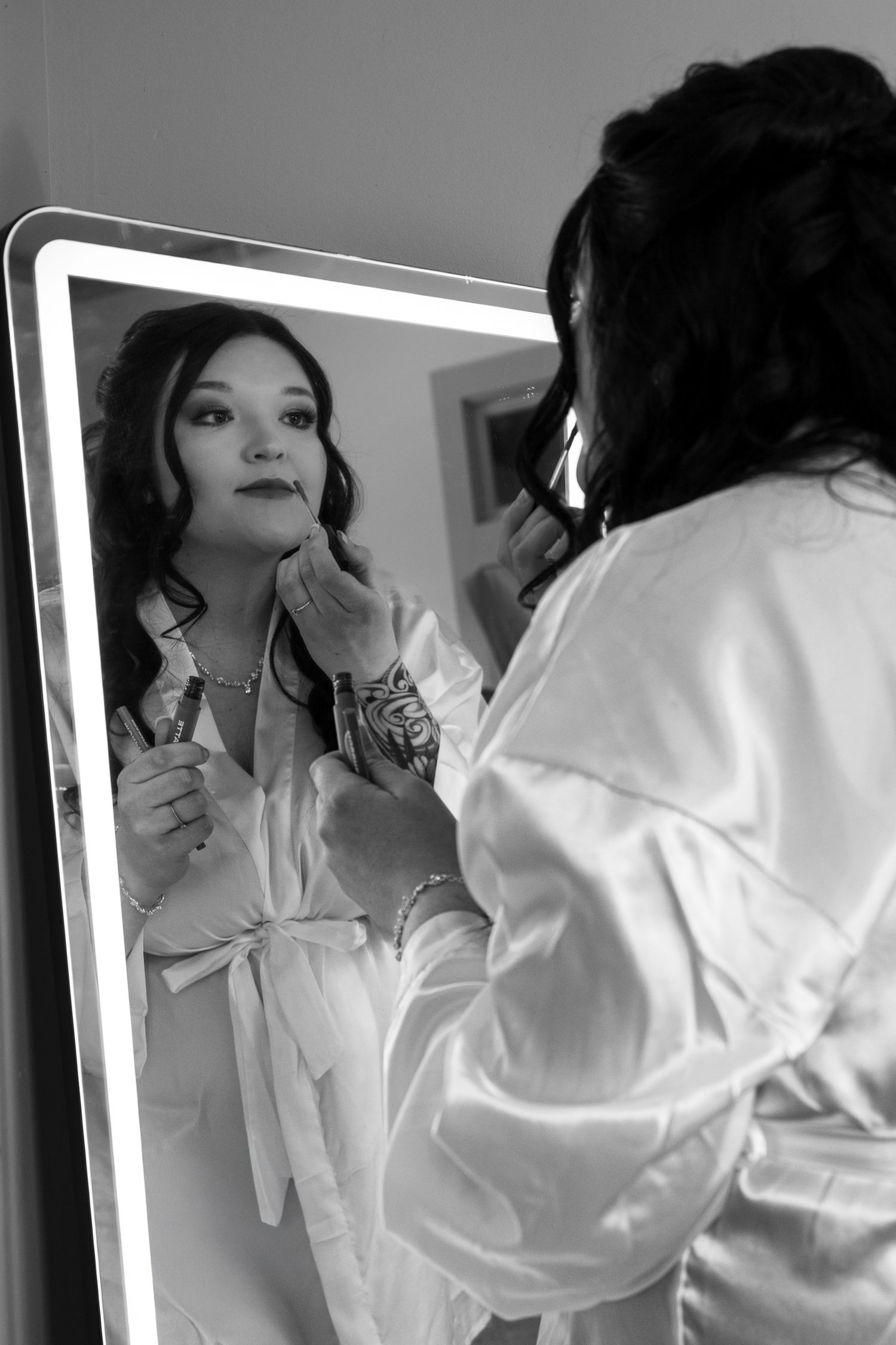 Bride applying lipstick in illuminated mirror wearing satin robe, black and white wedding preparation photo.