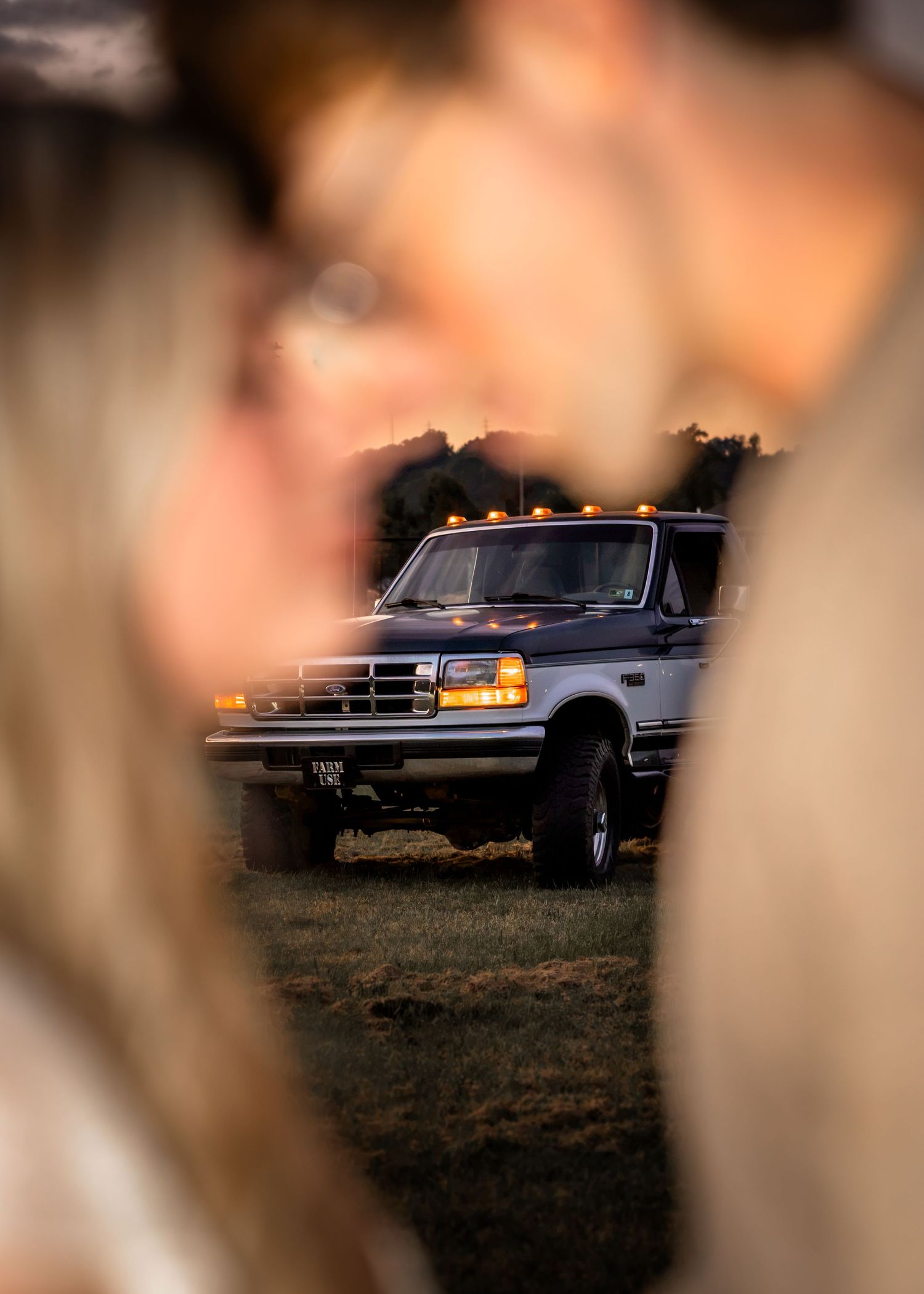 A vintage Ford Bronco SUV with amber roof lights glows in golden hour light, framed by two blurred figures in the foreground.