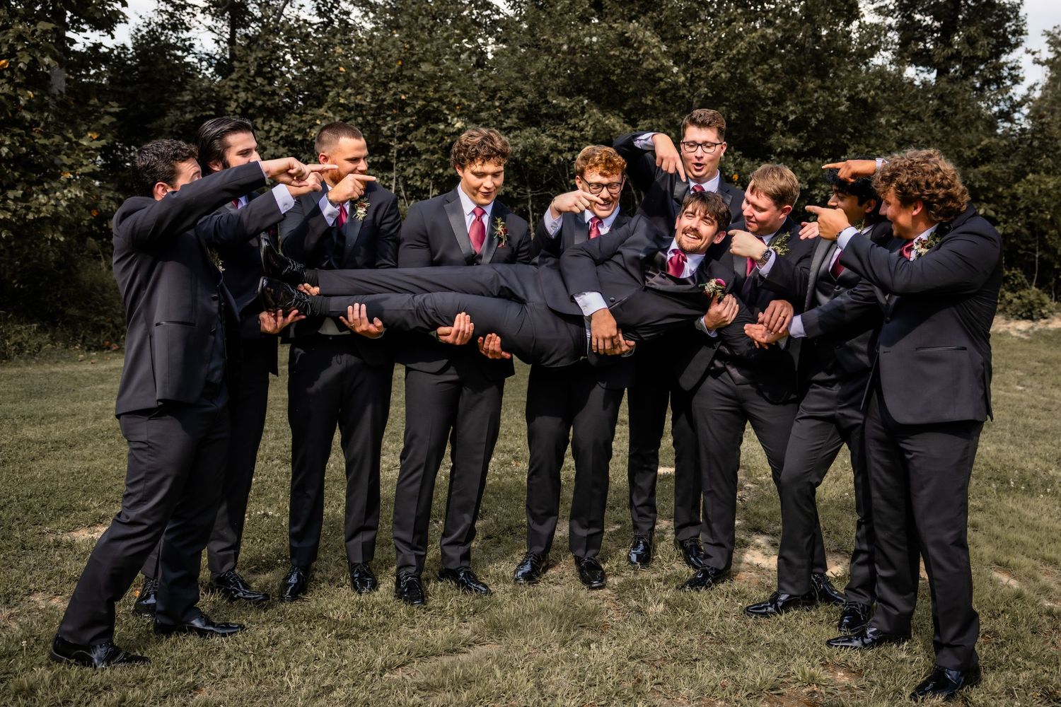 Groomsmen in dark suits lifting groom during fun outdoor wedding party photo on green lawn.