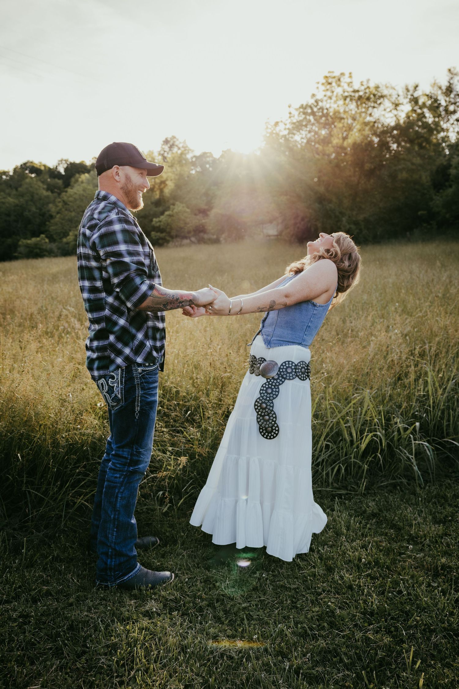 Couple holding hands and dancing in a golden sunlit field at sunset, man in plaid shirt and woman in white skirt.